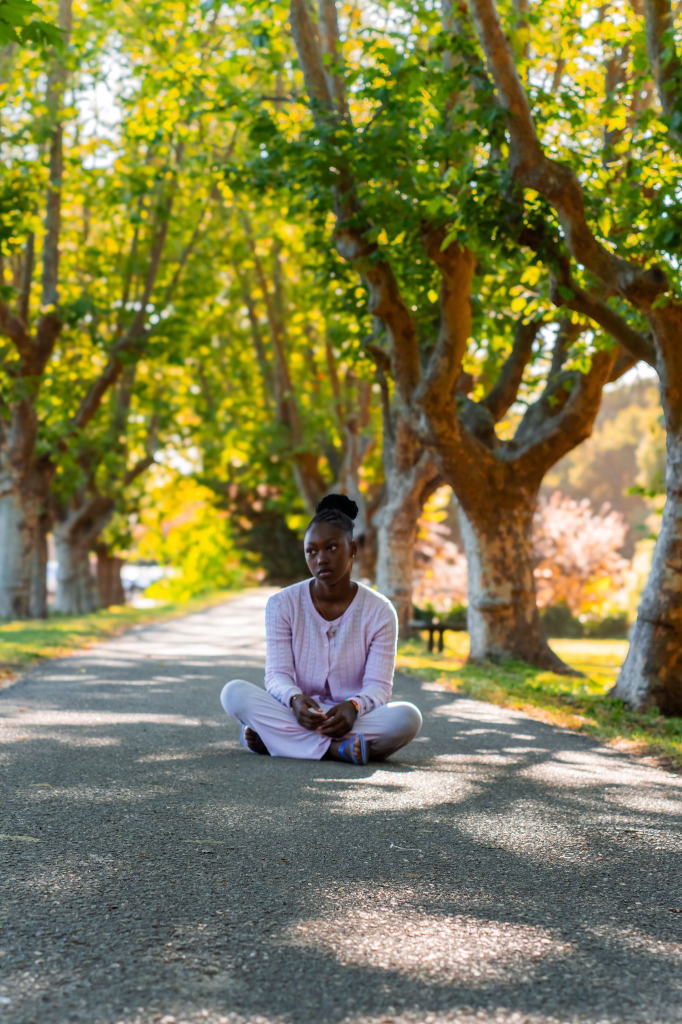 A young Black woman sits cross-legged on a tree-lined park path, wearing a pink outfit, surrounded by lush green foliage.