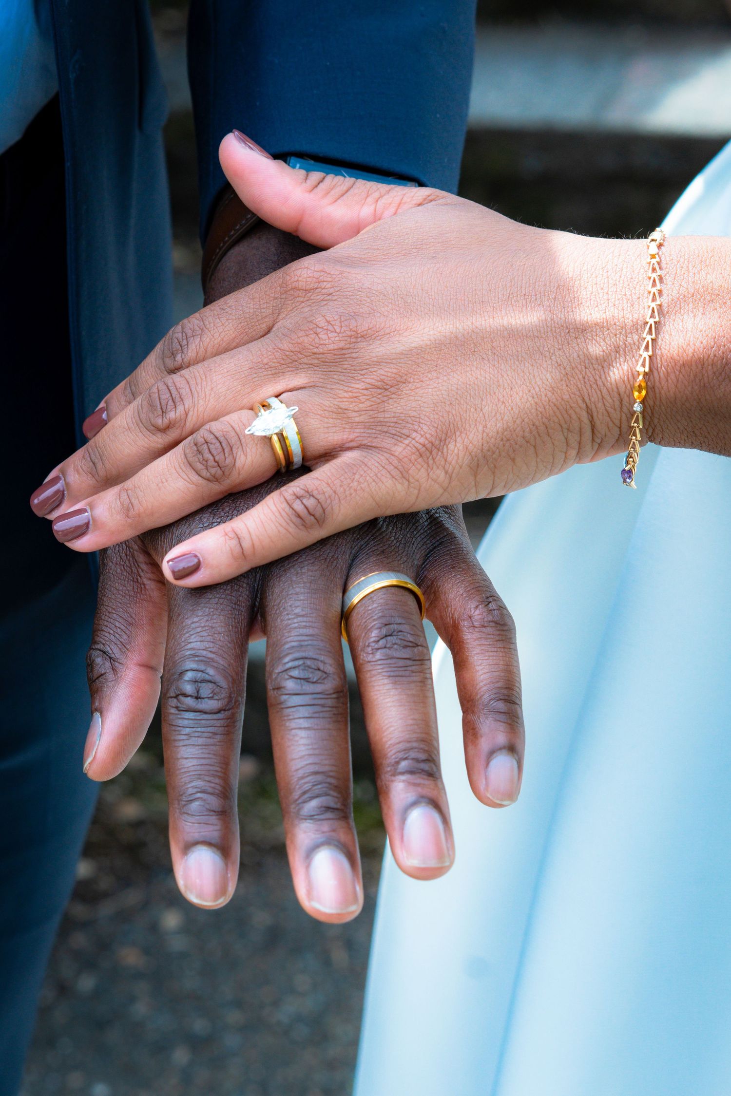 Close-up of newlywed hands showing wedding rings, the bride wearing a gold diamond ring and bracelet over a white dress.