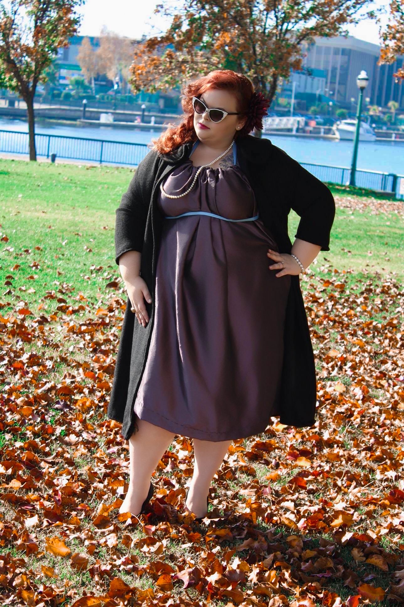Plus-size woman in purple dress and black cardigan standing in autumn leaves by a waterfront park on a sunny day.