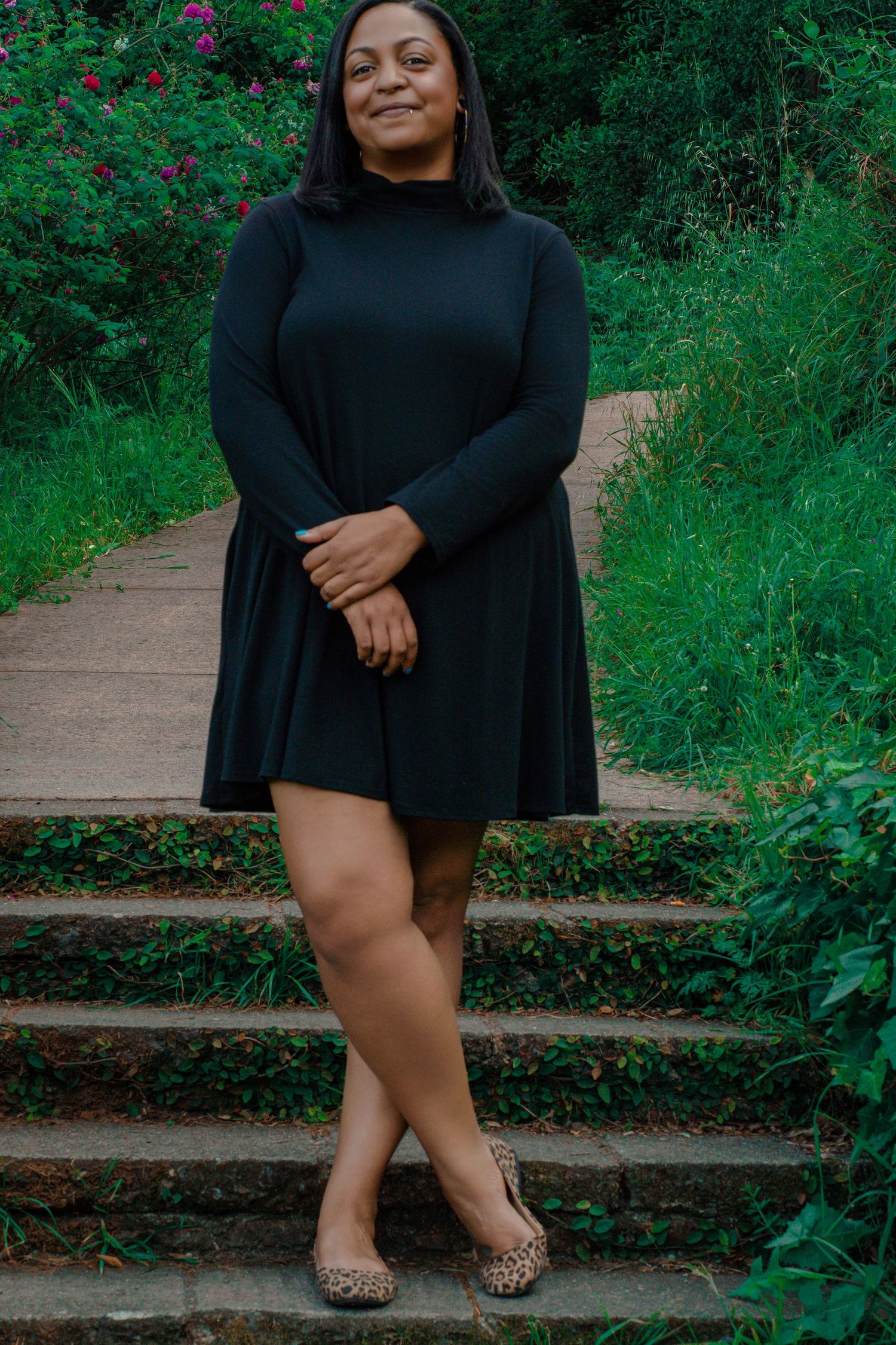 A Black woman in a black long-sleeve mini dress stands on stone steps with green foliage in the background.