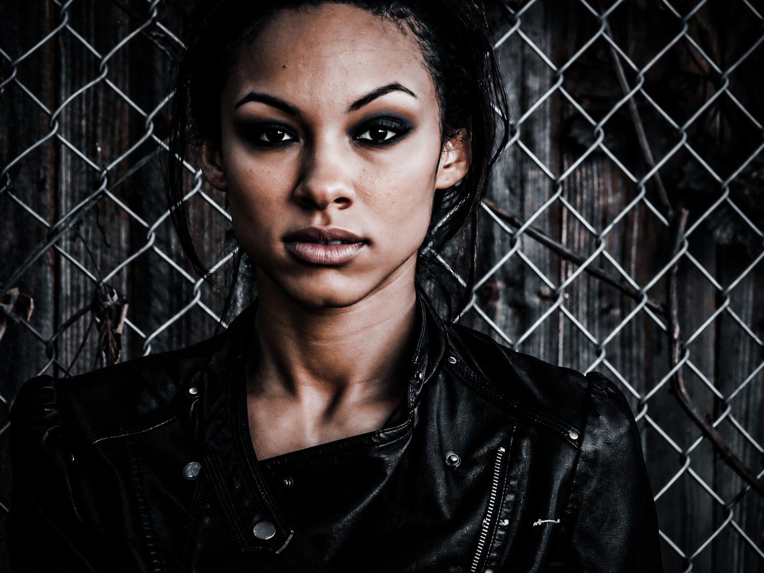 Young woman in black leather jacket posing against chain-link fence with dramatic dark lighting.