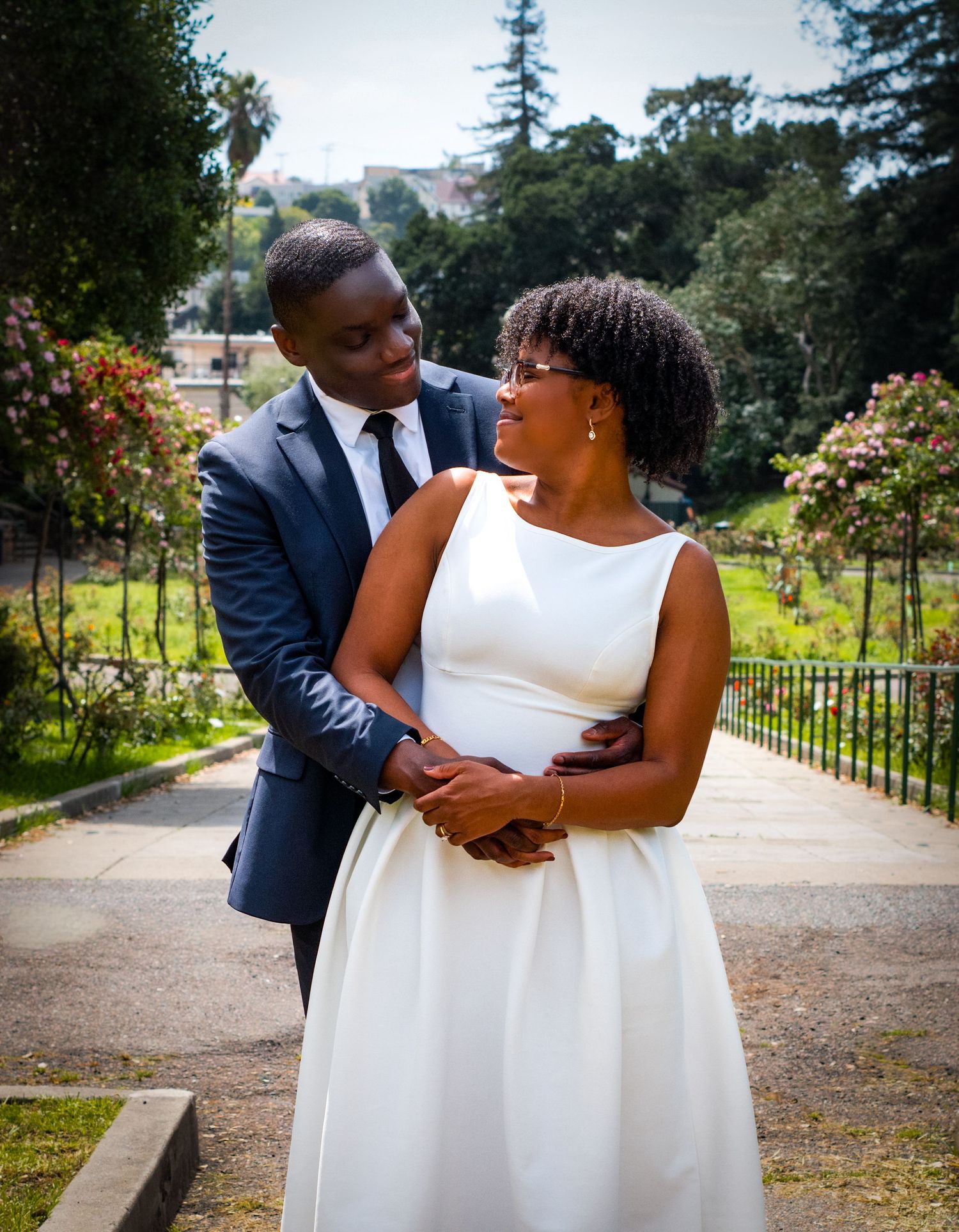 Couple in wedding attire embracing in a garden, woman in white dress and man in navy suit smiling at each other.