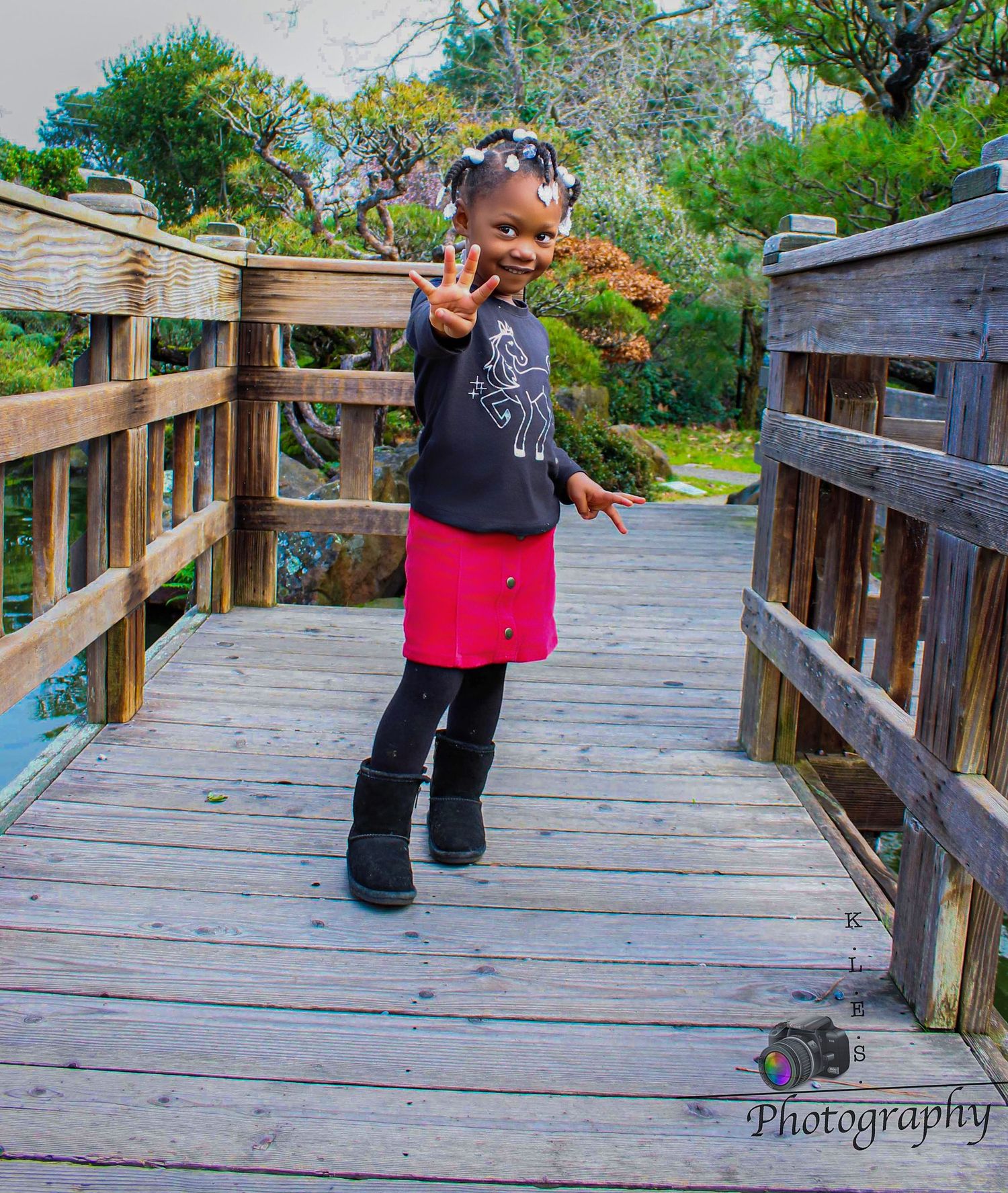 Happy young Black girl in pink skirt and black top making peace sign on a wooden bridge in a park.