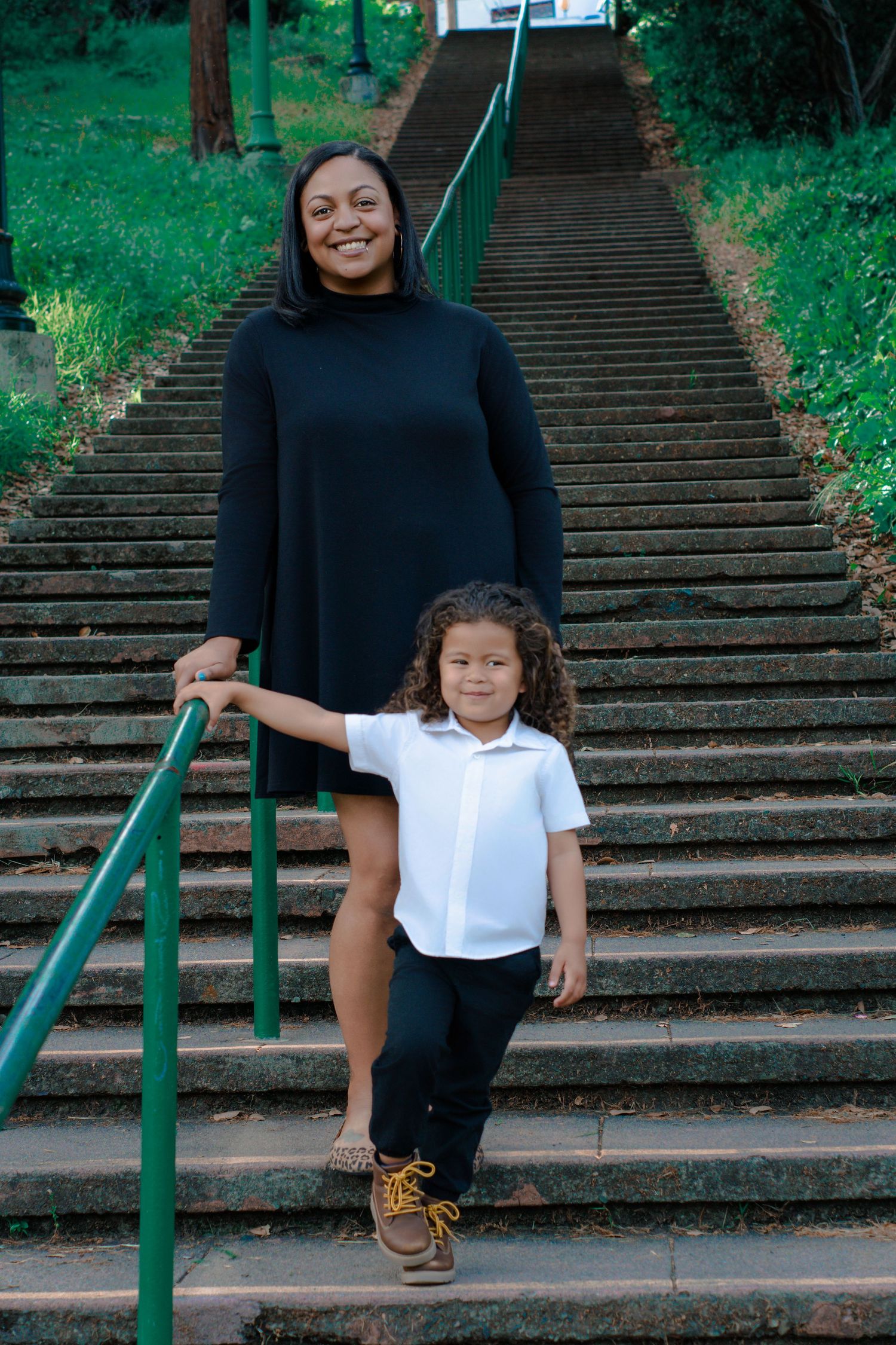 A Black woman in a black dress and a curly-haired child in a white polo pose on outdoor stone stairs.