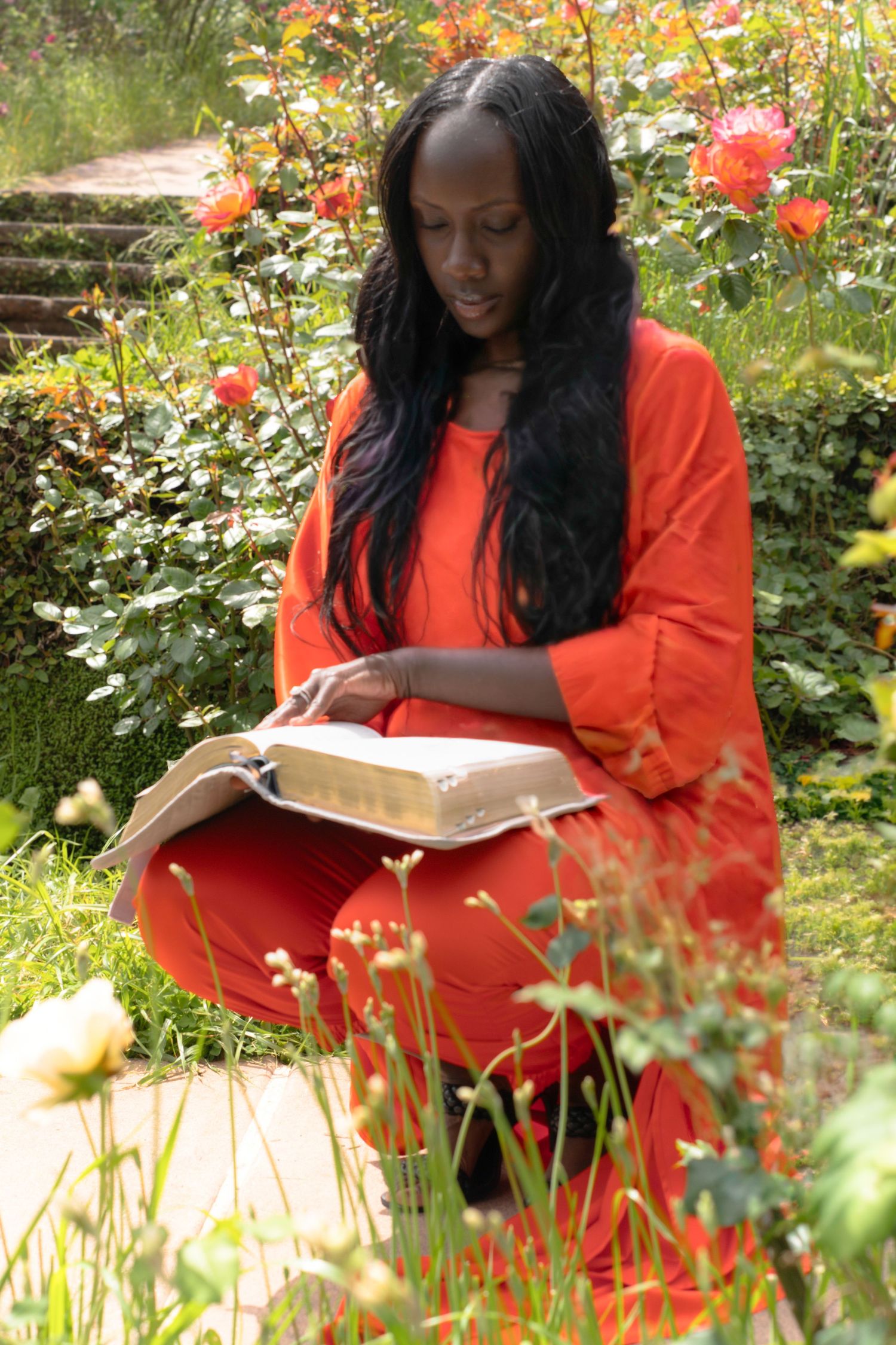 Black woman in orange outfit reading a book while sitting in a lush rose garden.