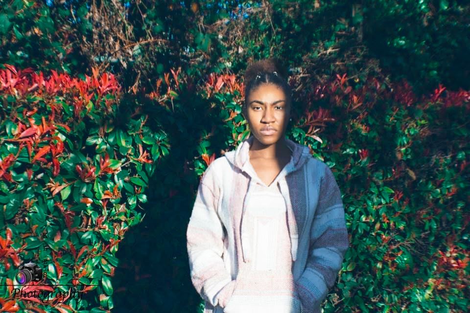 Young woman in gray hoodie stands confidently before vibrant red and green foliage backdrop.