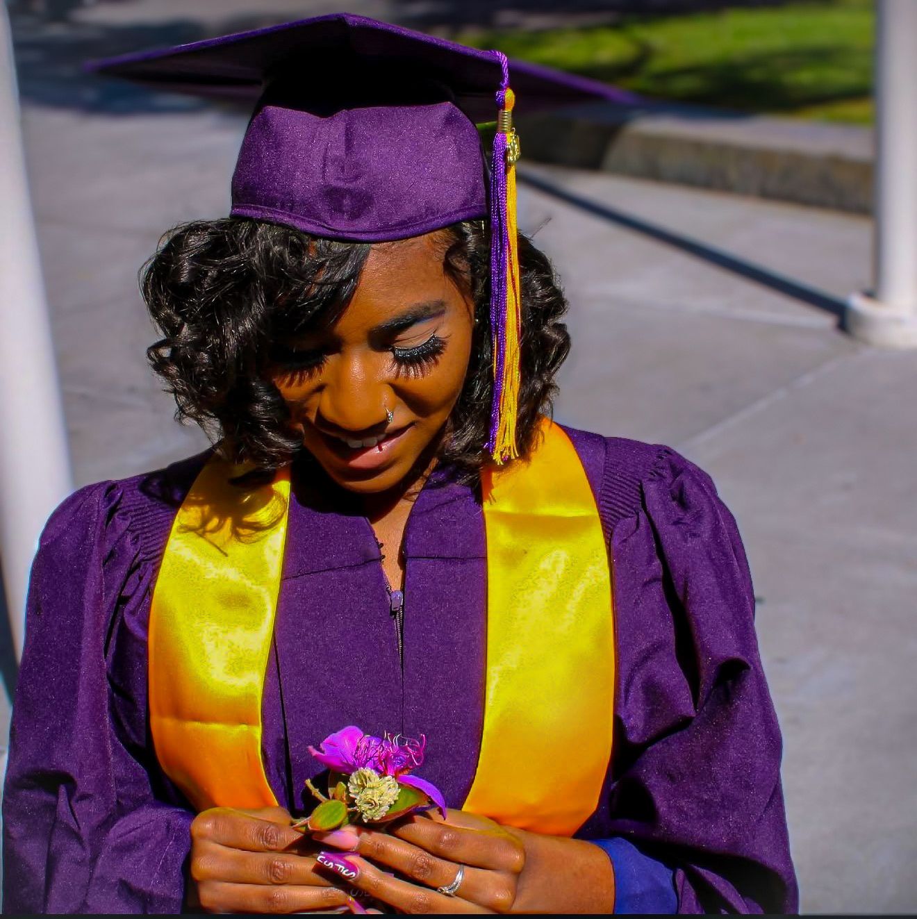 A young woman in purple graduation cap and gown with gold sash holds a purple flower corsage outdoors.