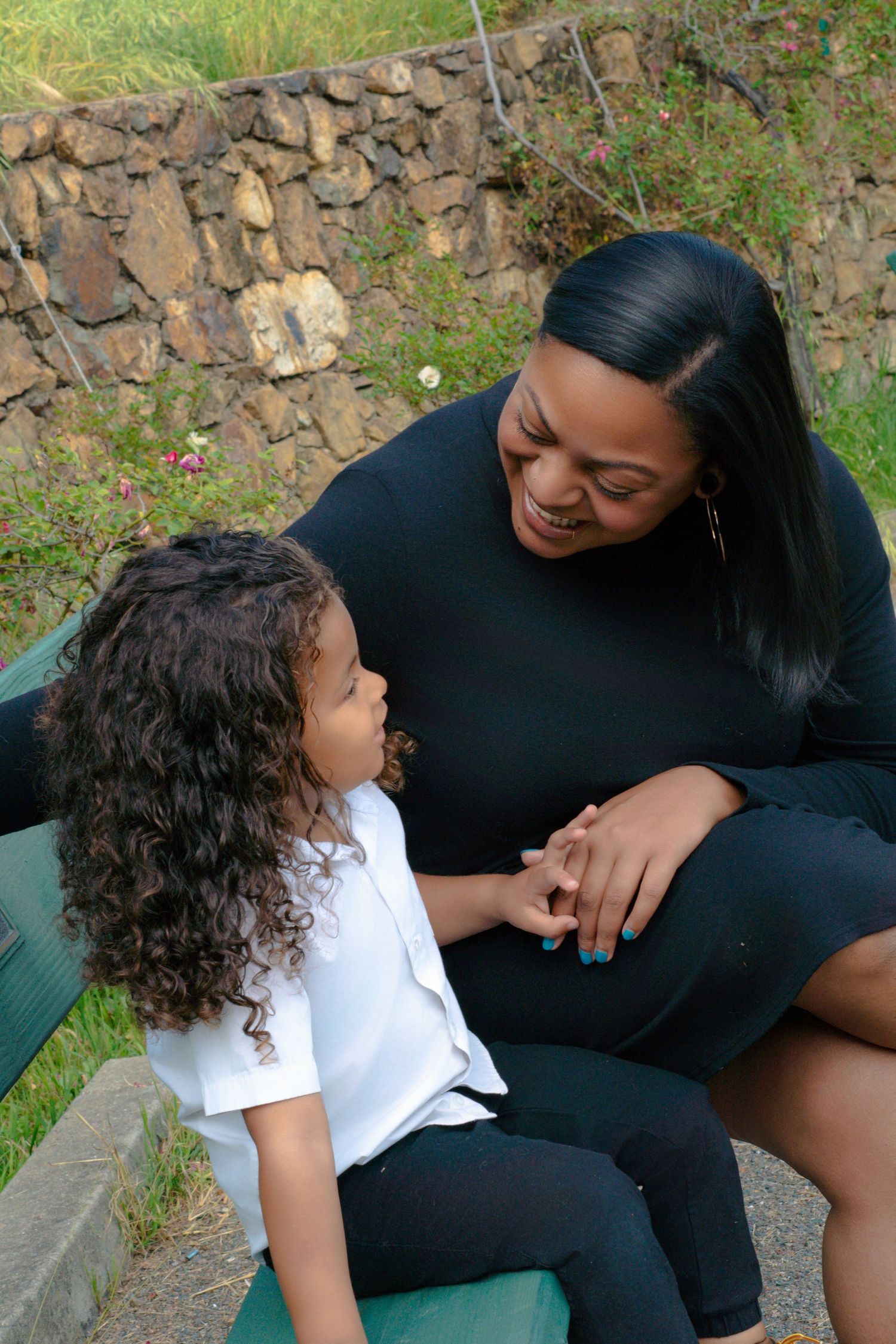 A Black woman in black smiles warmly at a curly-haired child in a white shirt while seated outdoors.
