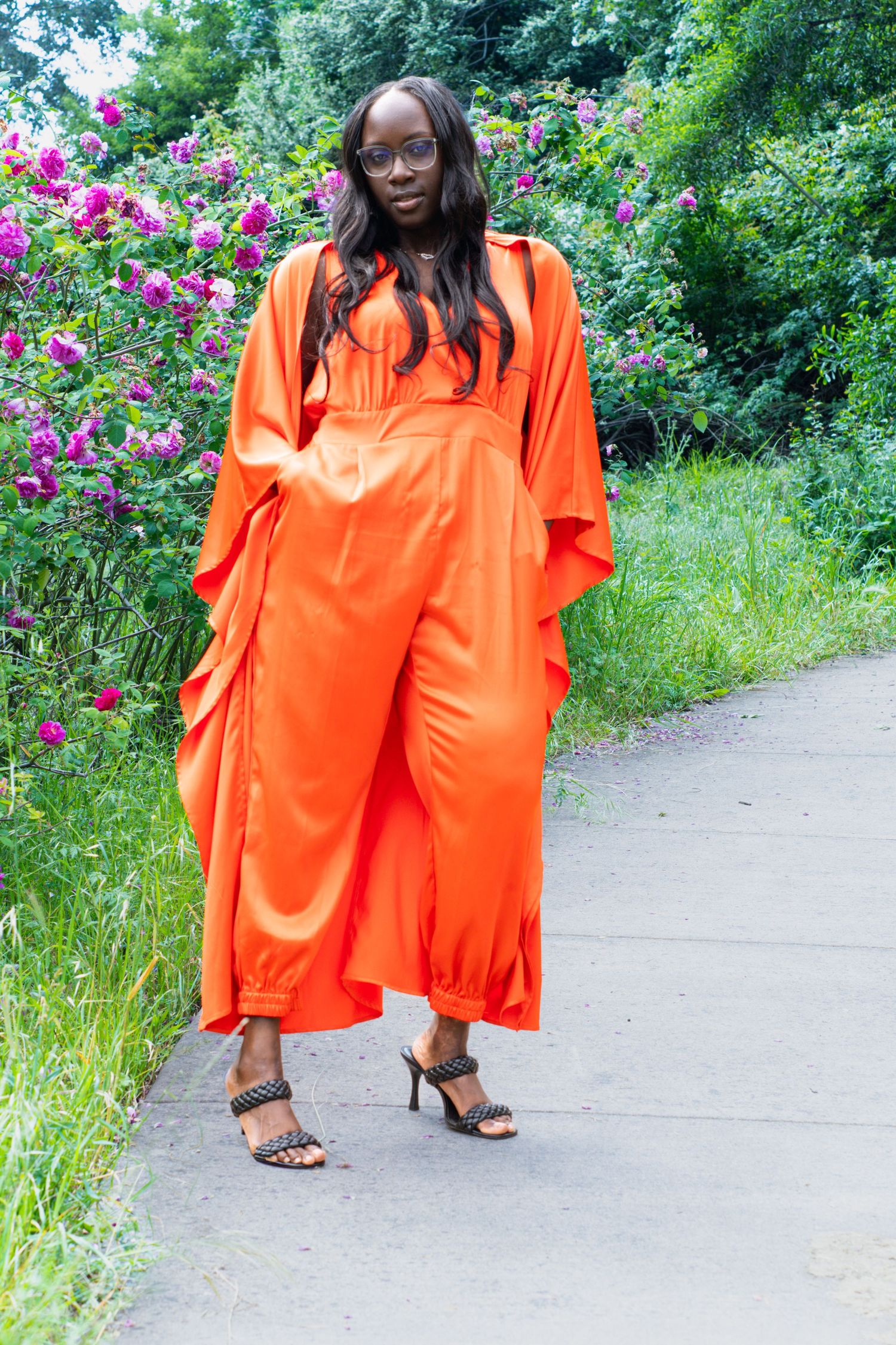 Black woman in bold orange jumpsuit and duster posing on path with pink flowers.