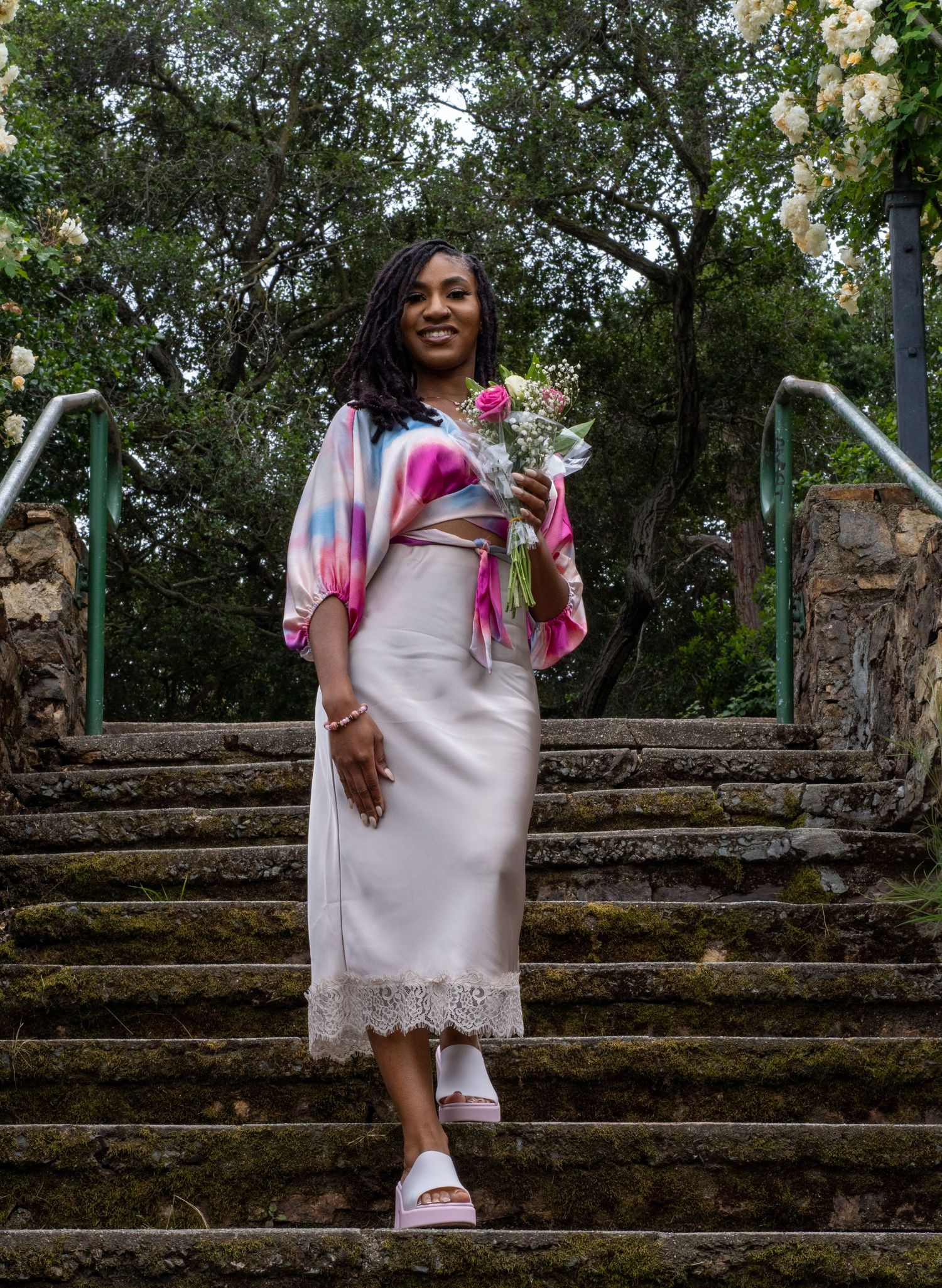 Woman in vibrant tie-dye top and cream lace skirt holding bouquet on moss-covered stone steps.