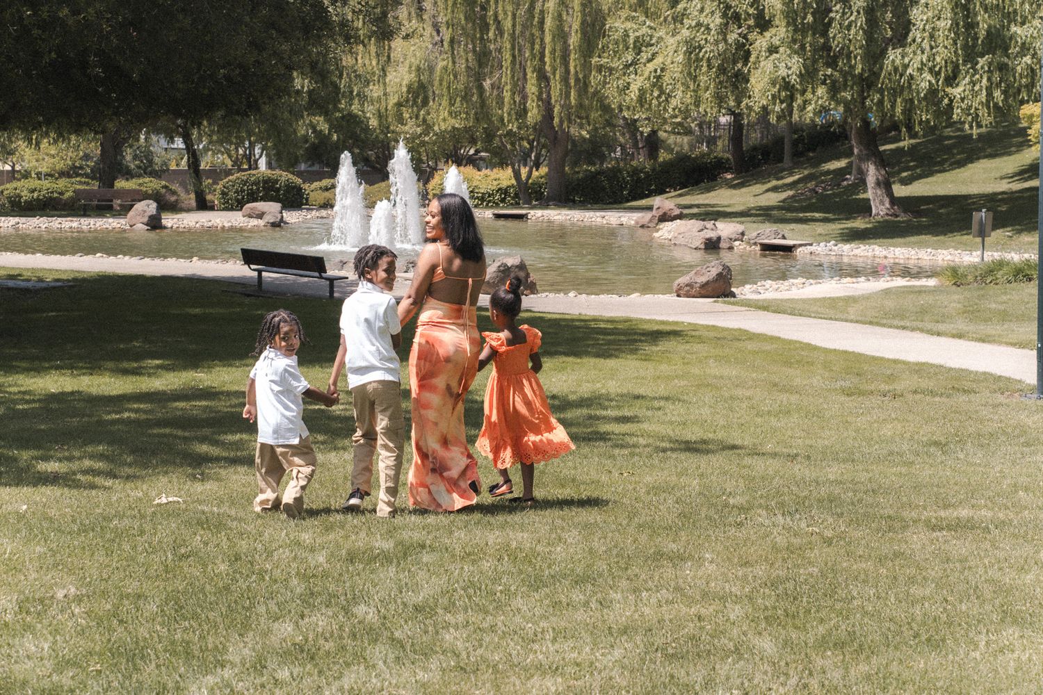 Woman in orange dress walks with three children in a sunny green park with fountain backdrop.