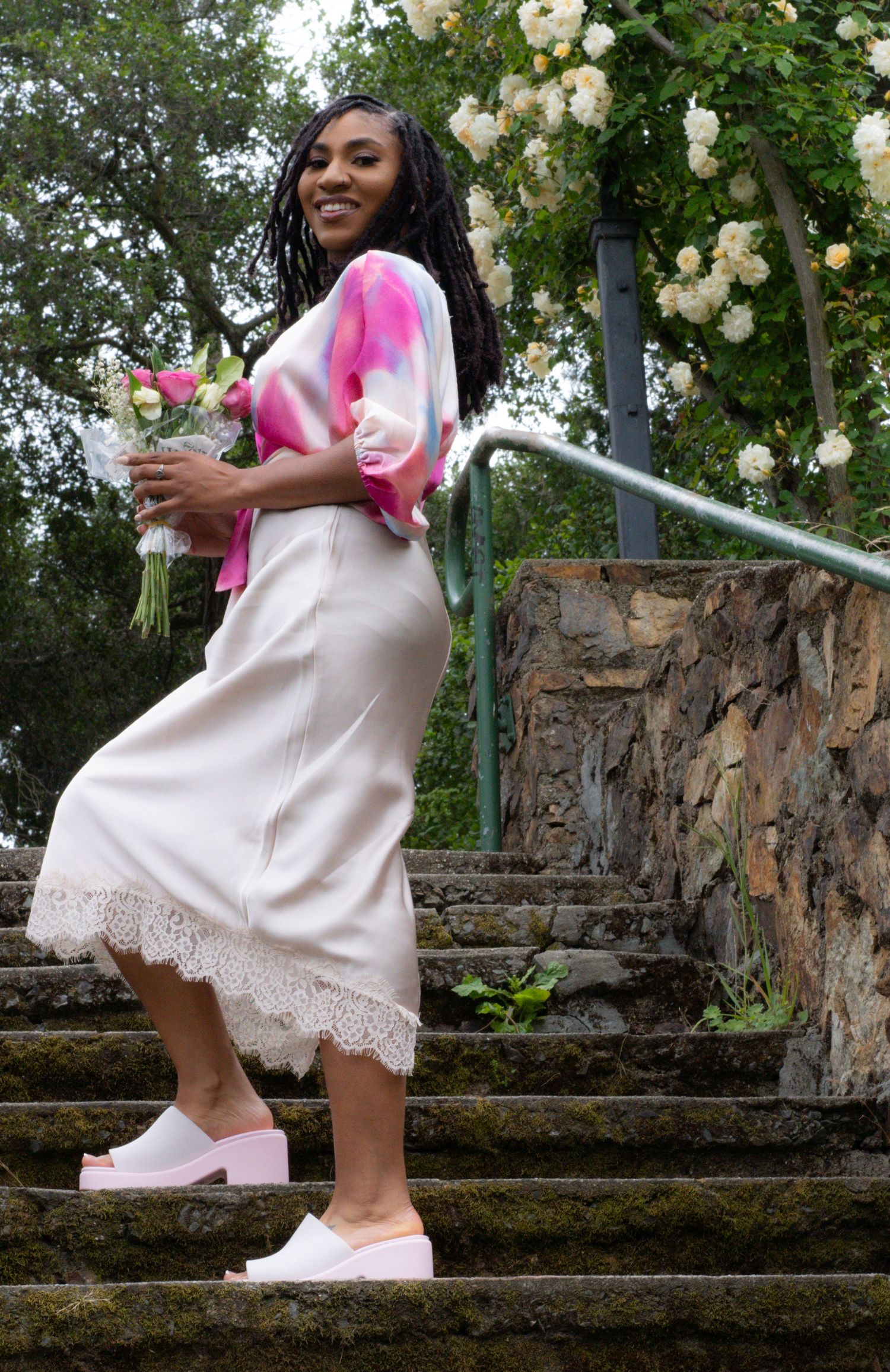 Smiling woman in pink tie-dye blouse and cream lace midi skirt walking up stone steps with flowers.