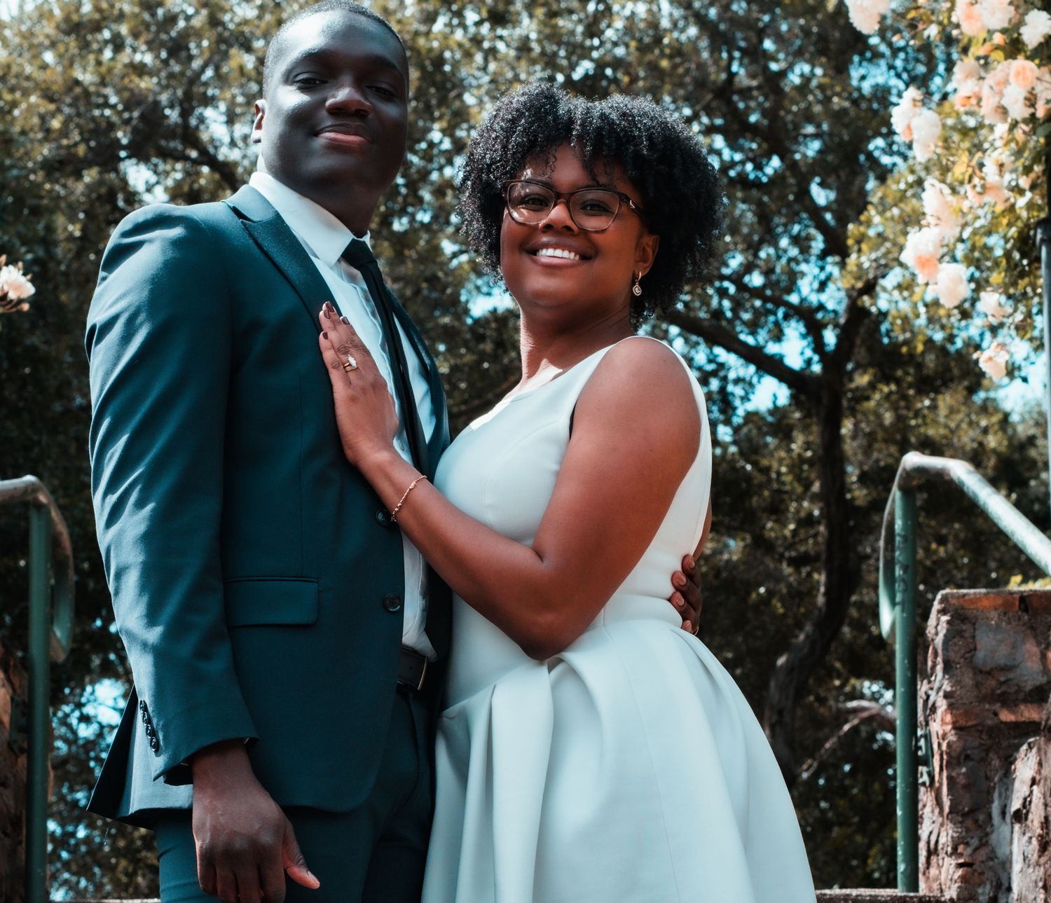 A couple in formal attire poses outdoors, man in teal suit, woman in light blue dress, smiling among trees.