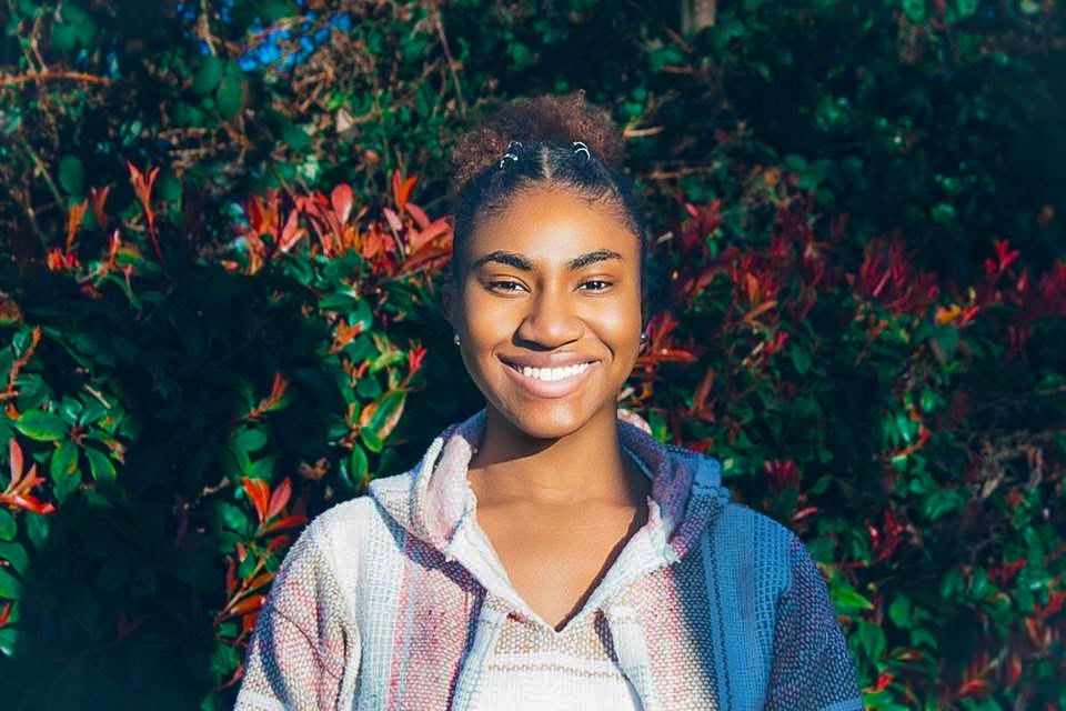 Smiling young Black woman with natural updo hairstyle standing outdoors in front of lush green and red foliage.