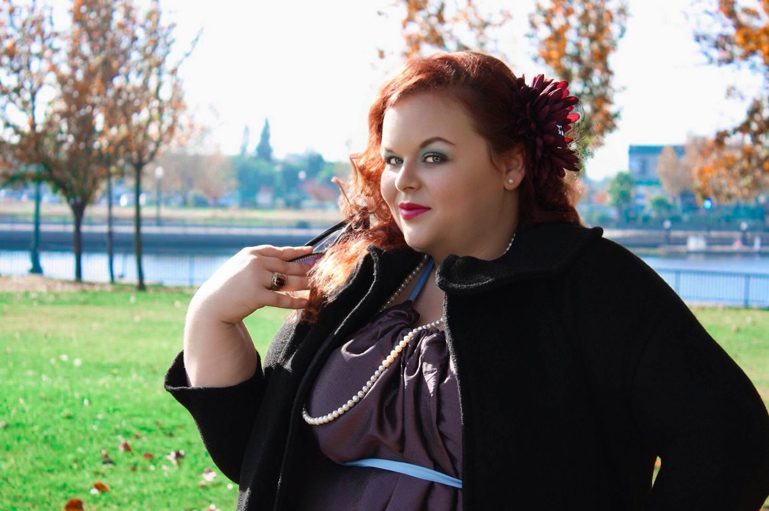 Plus-size woman with red hair and flower accessory wearing a black cape outdoors in a park during autumn.