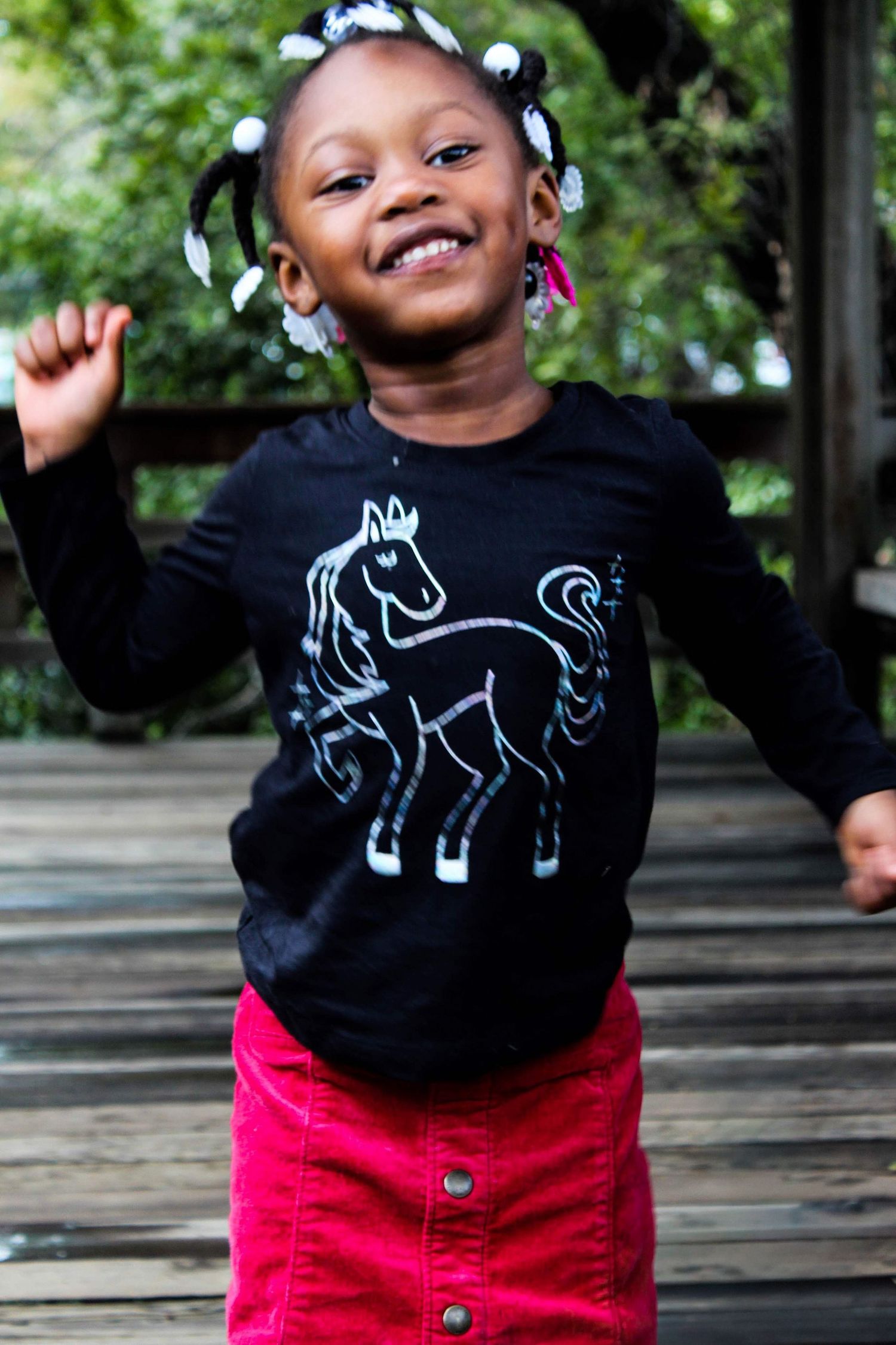 A joyful young Black girl in a black horse-print shirt and red skirt dances and smiles on a wooden deck outdoors.