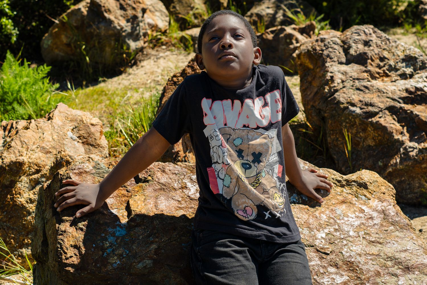 Young boy in graphic tee sitting on rocky terrain surrounded by lush greenery.