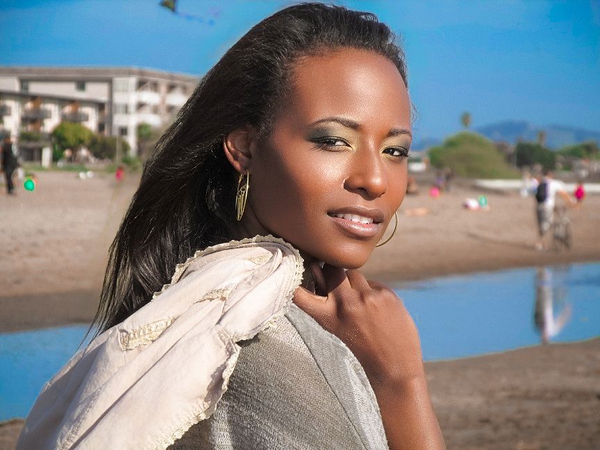 Elegant woman with long dark hair poses on a sunny beach wearing a white scarf, with buildings visible behind her.