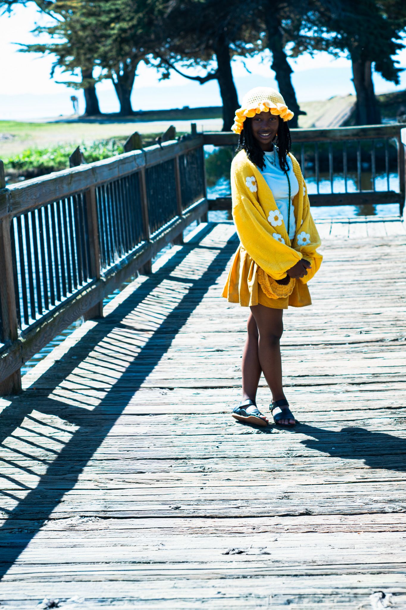 Young woman in yellow cardigan and skirt with floral bucket hat posing on a wooden boardwalk.