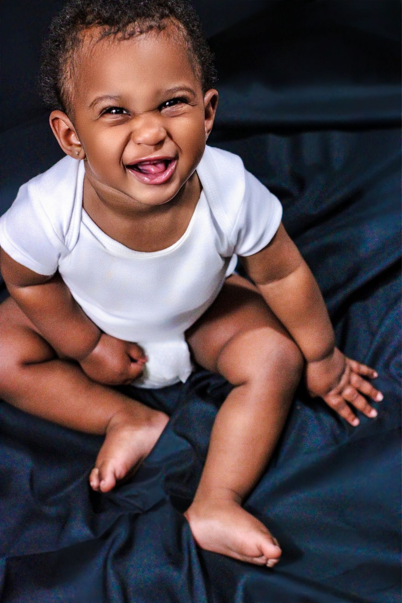 Smiling African American baby in white onesie sitting on dark fabric, laughing joyfully with bare feet.