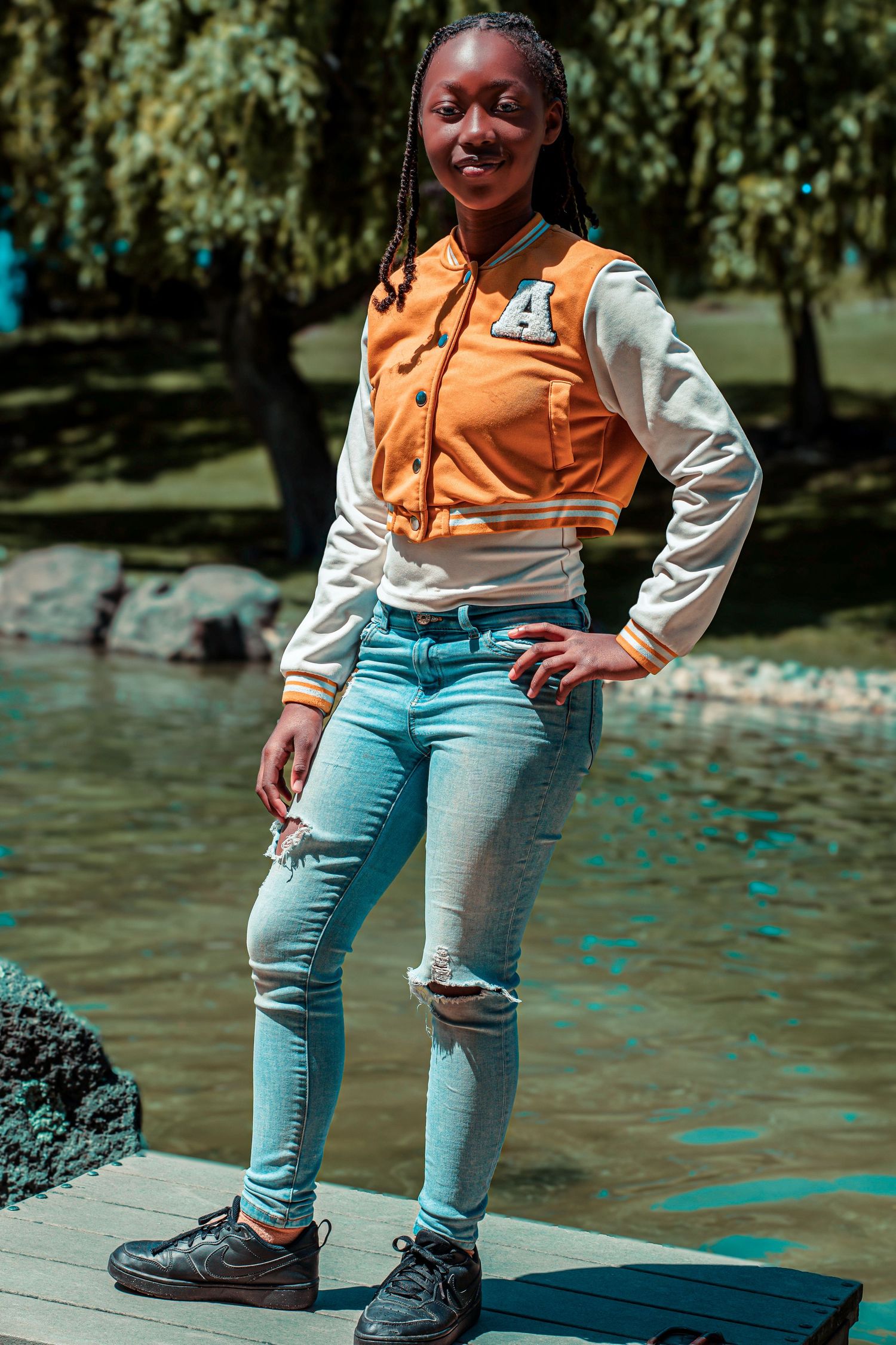 Young woman in orange varsity jacket and ripped jeans posing confidently by a scenic pond in a park.
