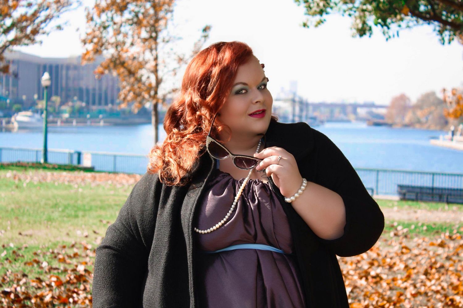 Plus-size woman in purple dress and black cardigan poses outdoors near waterfront with autumn foliage.
