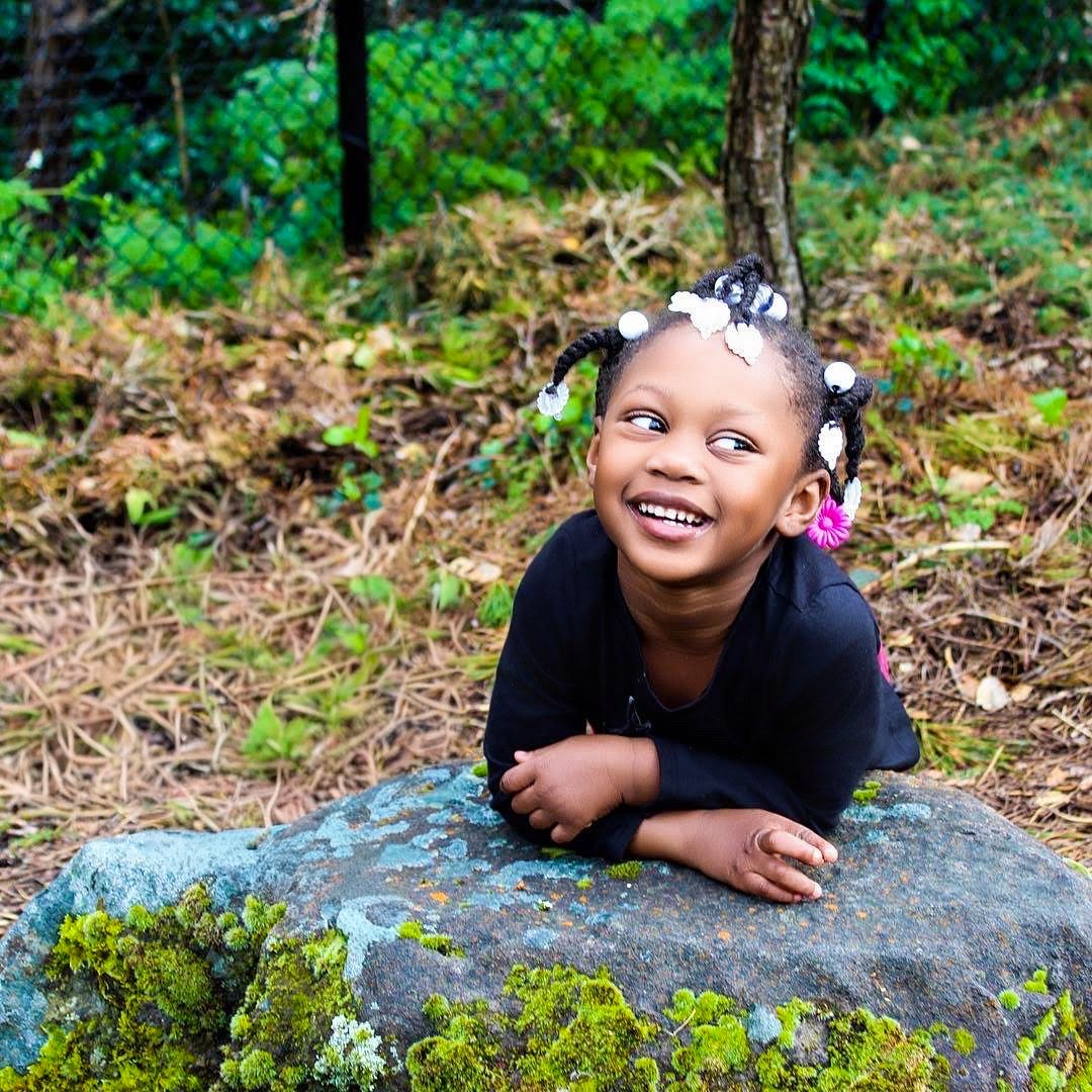 Joyful young girl with pigtails leaning on a moss-covered rock, smiling outdoors in a lush green setting.