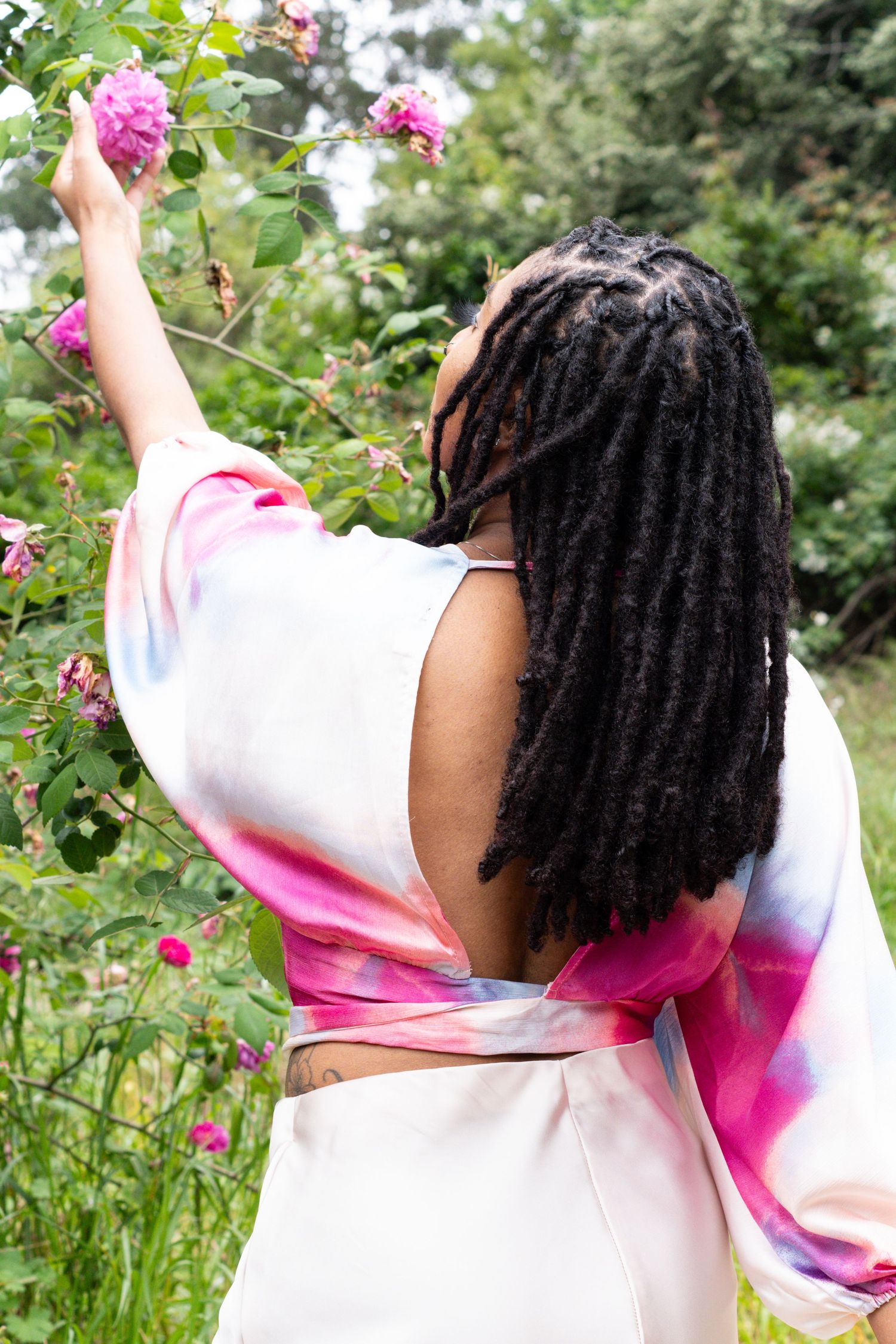Back view of woman with locs in open-back tie-dye top reaching for pink roses in garden.