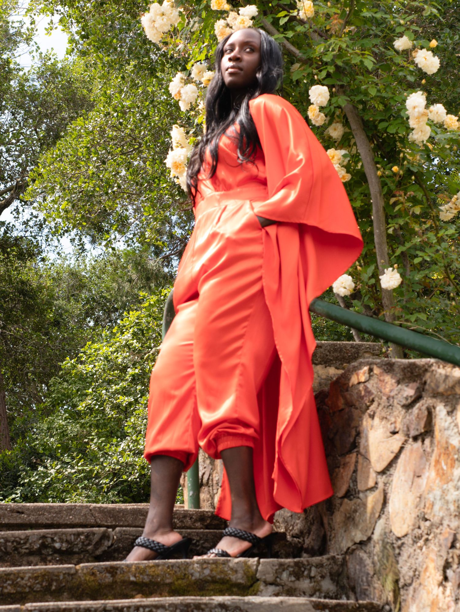 Tall Black woman in flowing orange satin dress standing on stone steps among roses.