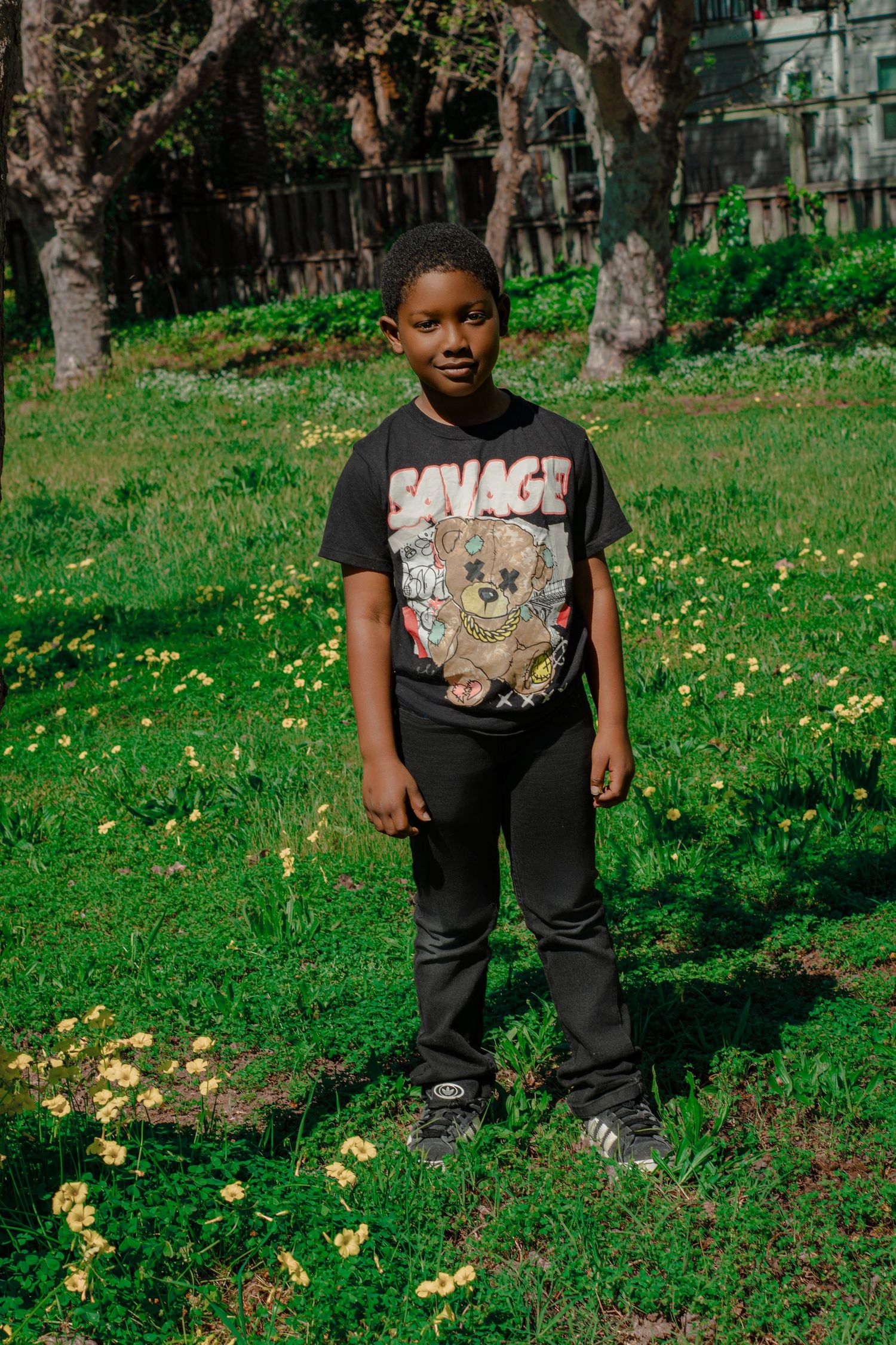 Young boy in a black Savage graphic tee stands in a sunny green field with yellow wildflowers and trees.