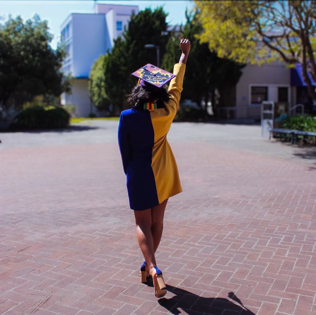 Graduate in yellow gown raises fist in celebration on brick campus plaza, wearing decorated mortarboard cap.