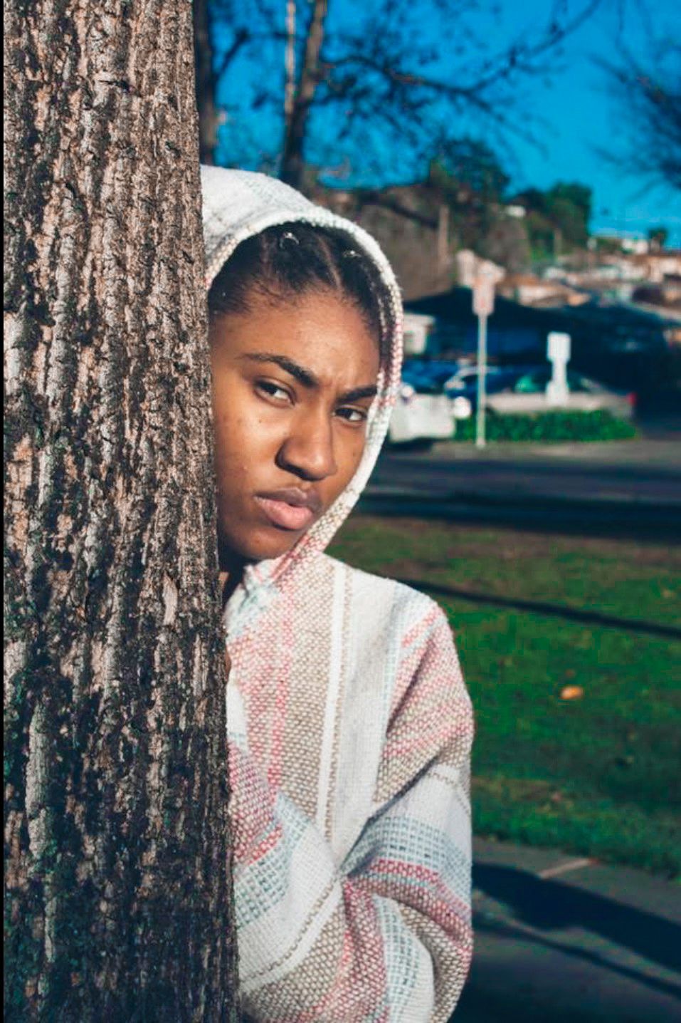 Young woman in a hooded sweater leaning against a tree trunk outdoors on a sunny day.