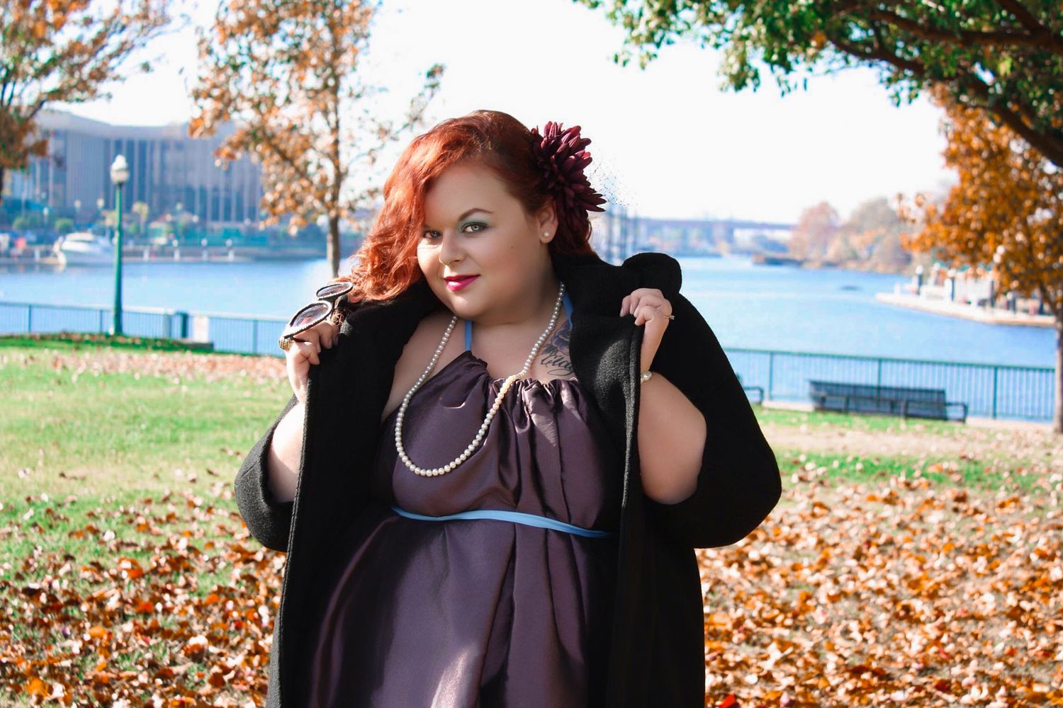 Plus-size woman in purple dress and black coat poses outdoors among autumn leaves near a river.