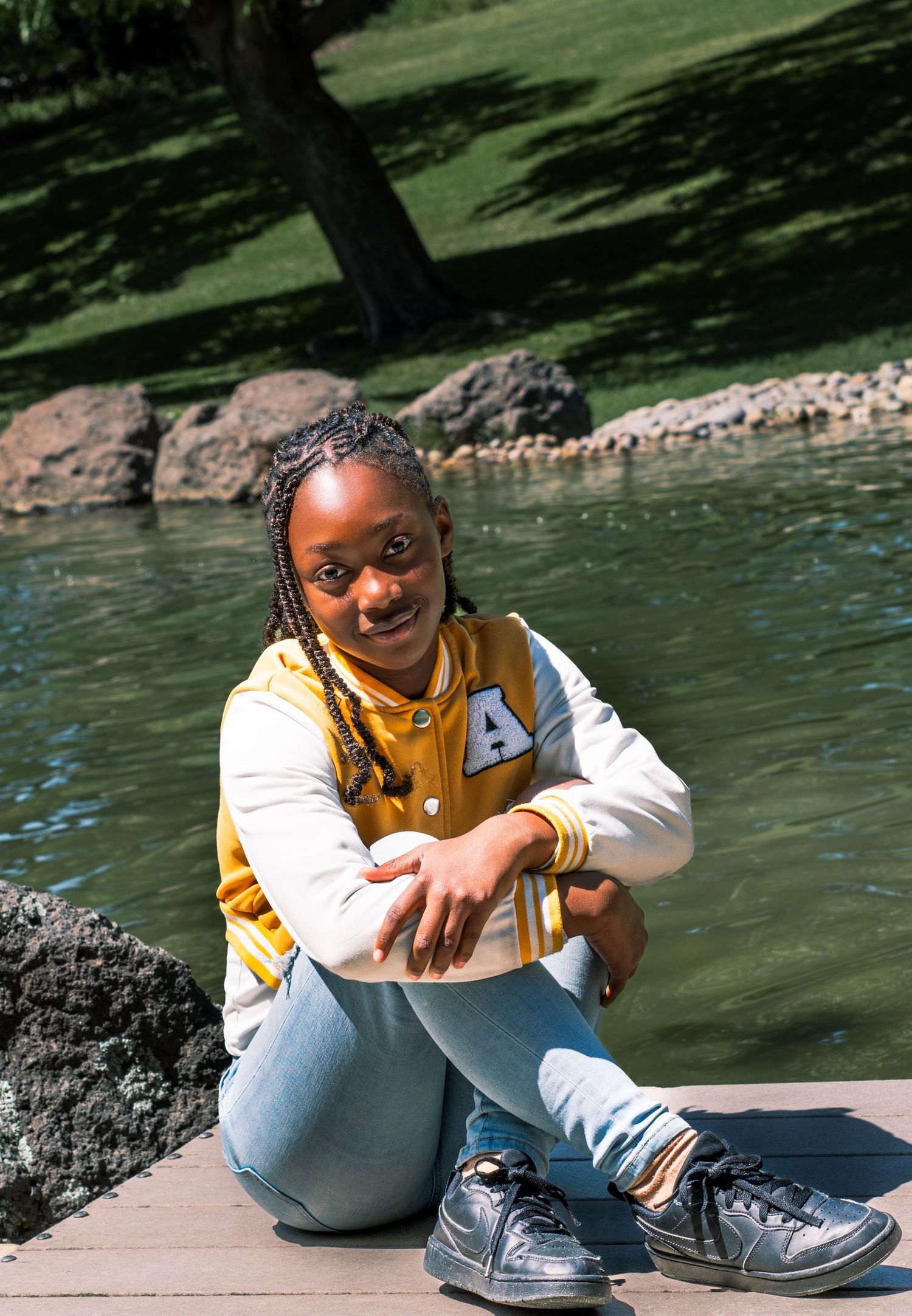 Young girl in yellow varsity jacket and jeans sits smiling by a serene pond in a sunny park.