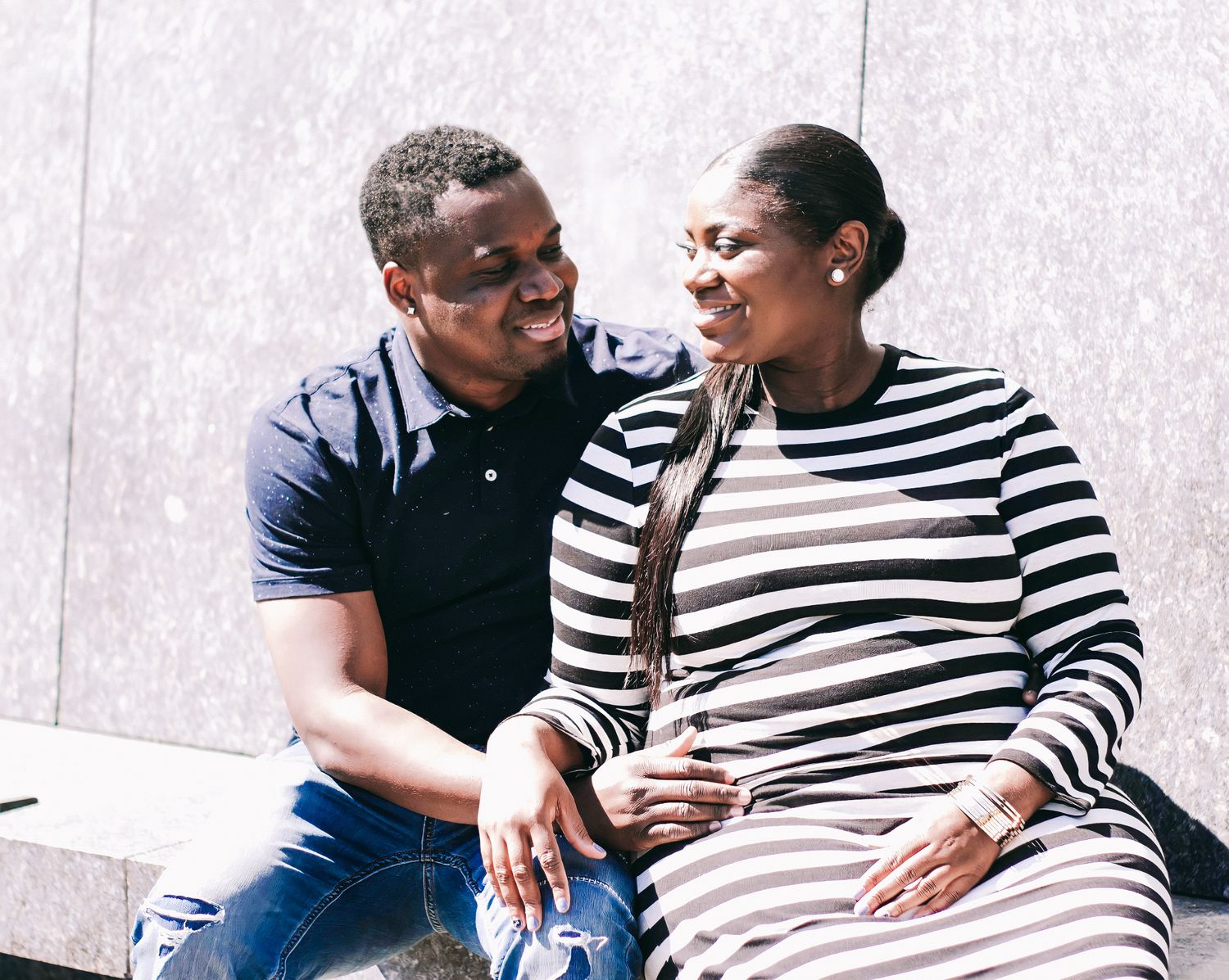 A happy couple sitting together outdoors, smiling at each other, woman wearing striped dress, man in navy polo shirt.