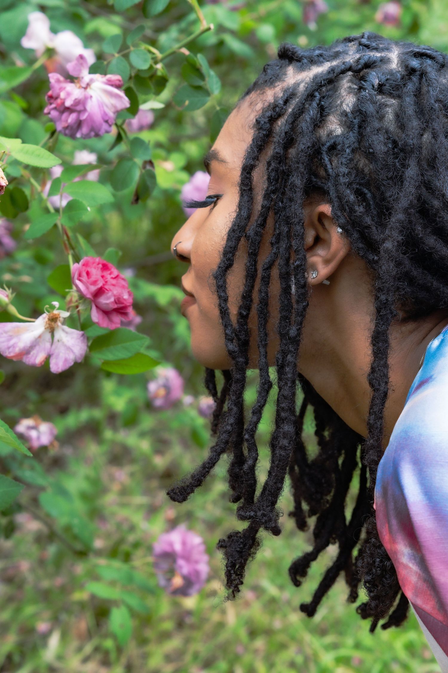 Side profile of woman with locs smelling pink roses on bush in lush green garden.