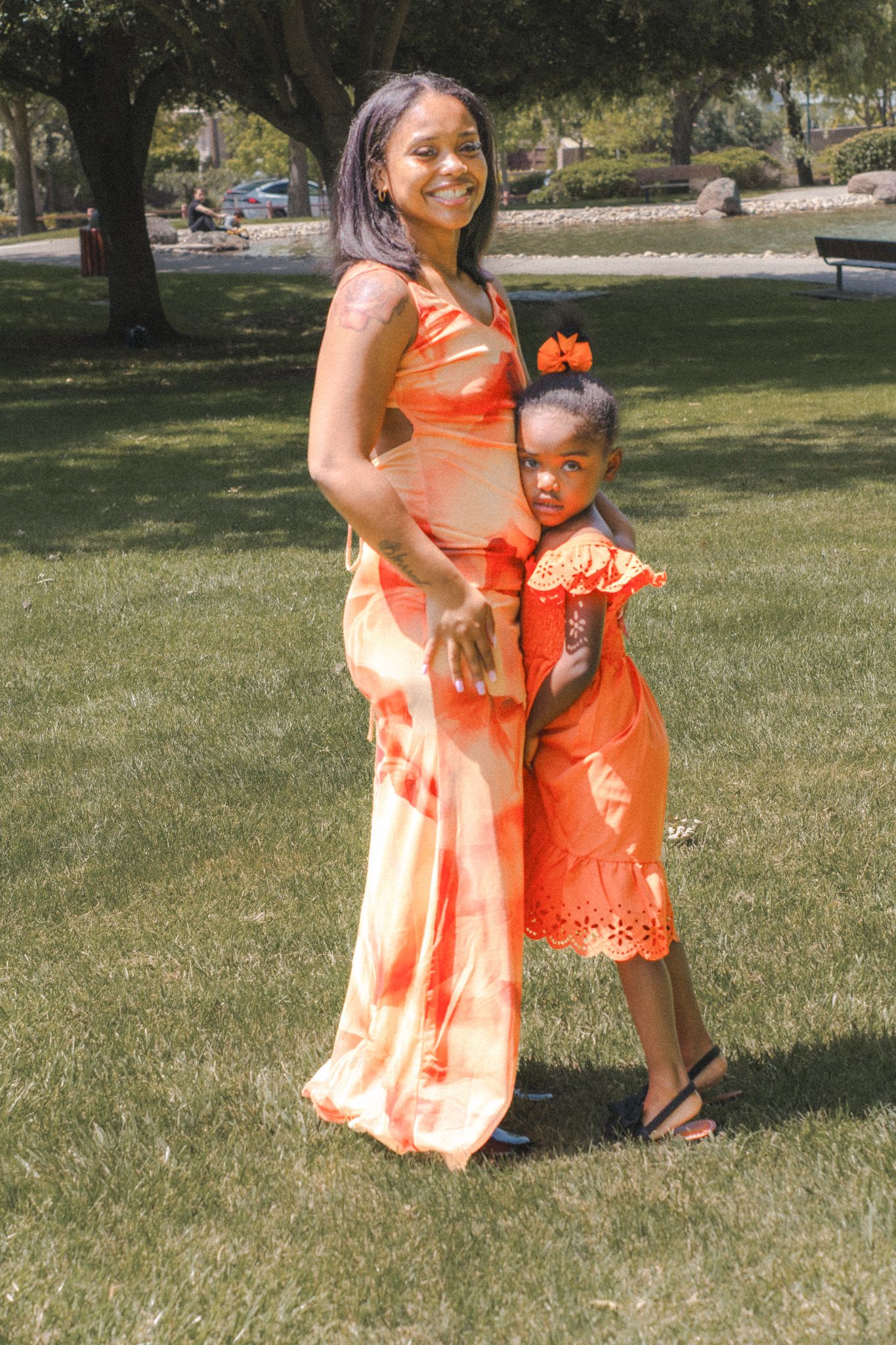 Smiling Black mother in orange tie-dye dress hugging her young daughter in a park.