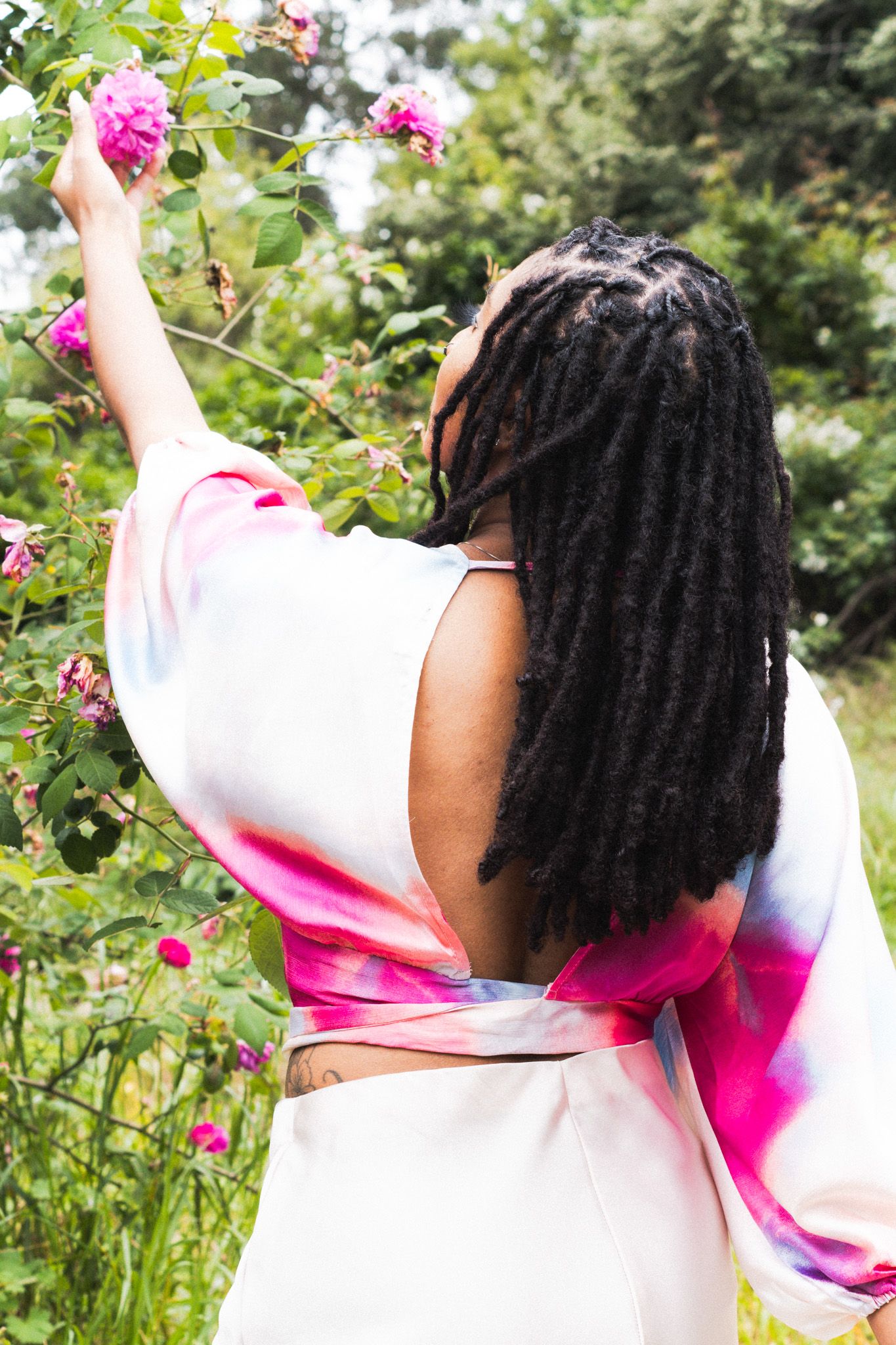 Woman with dreadlocks in a pink and white tie-dye top reaching up to touch pink roses in a garden.