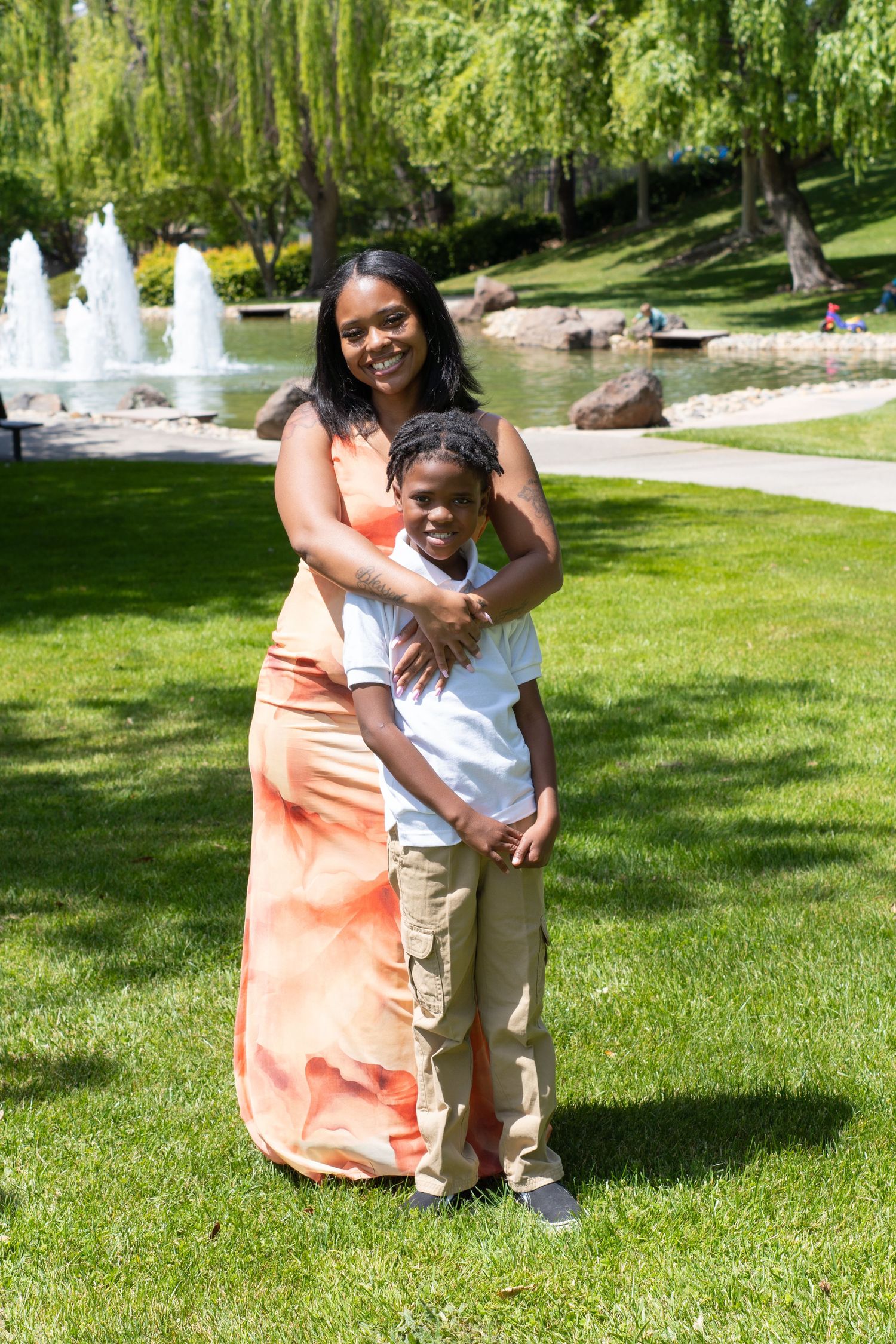 A Black woman in an orange maxi dress hugs a young boy in a white shirt in a sunny park near a fountain.