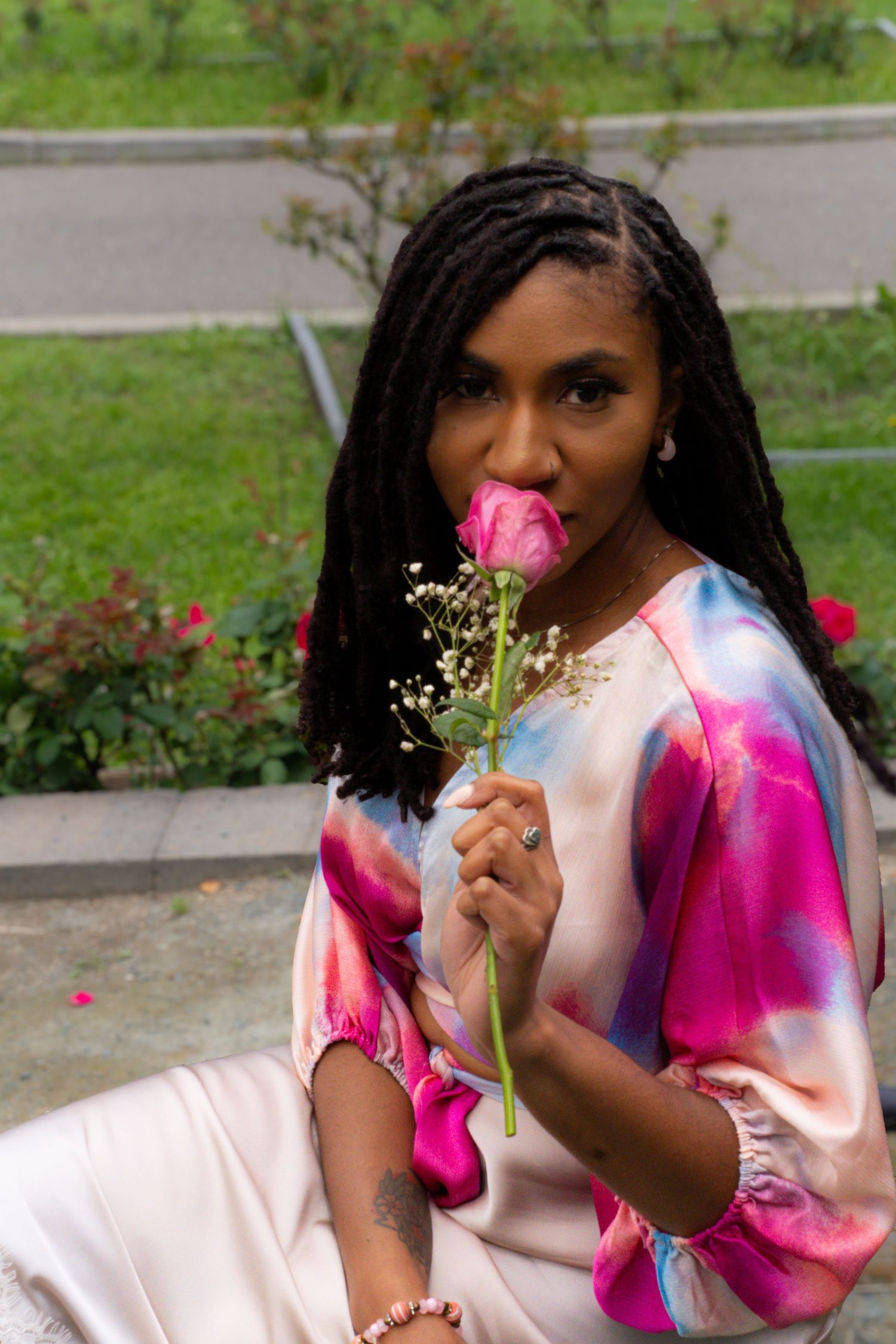 Black woman with braids smelling a pink rose in a colorful tie-dye top outdoors.