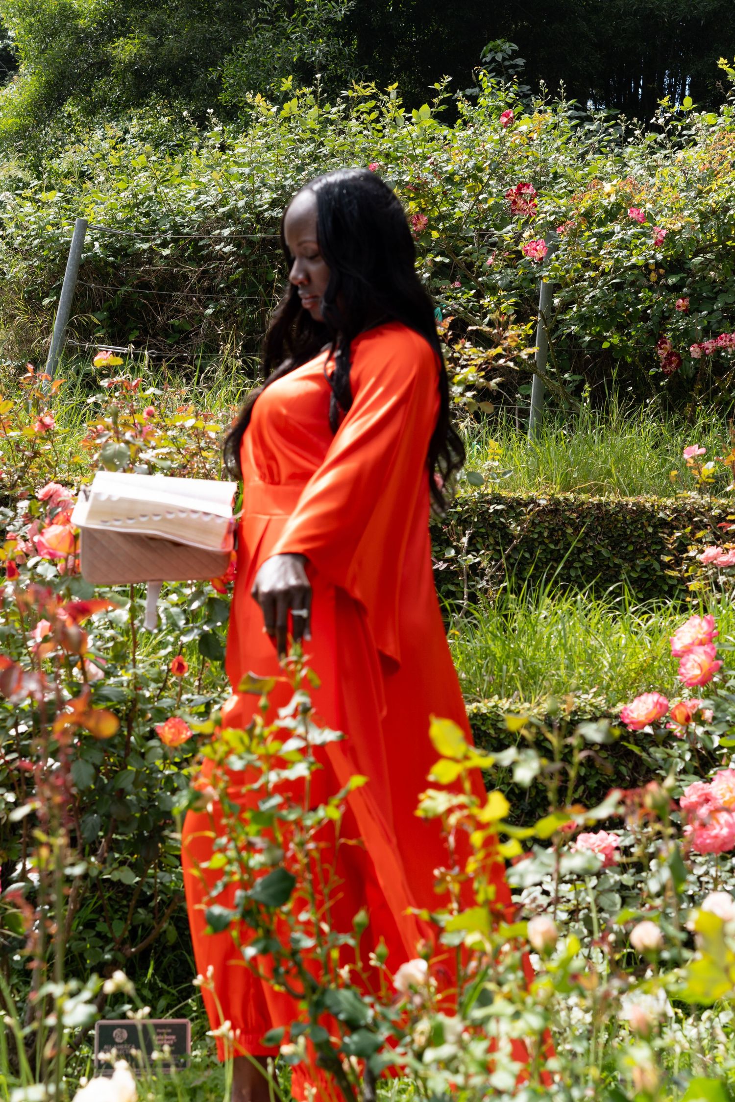Black woman in orange dress holding a book while walking through a vibrant rose garden.