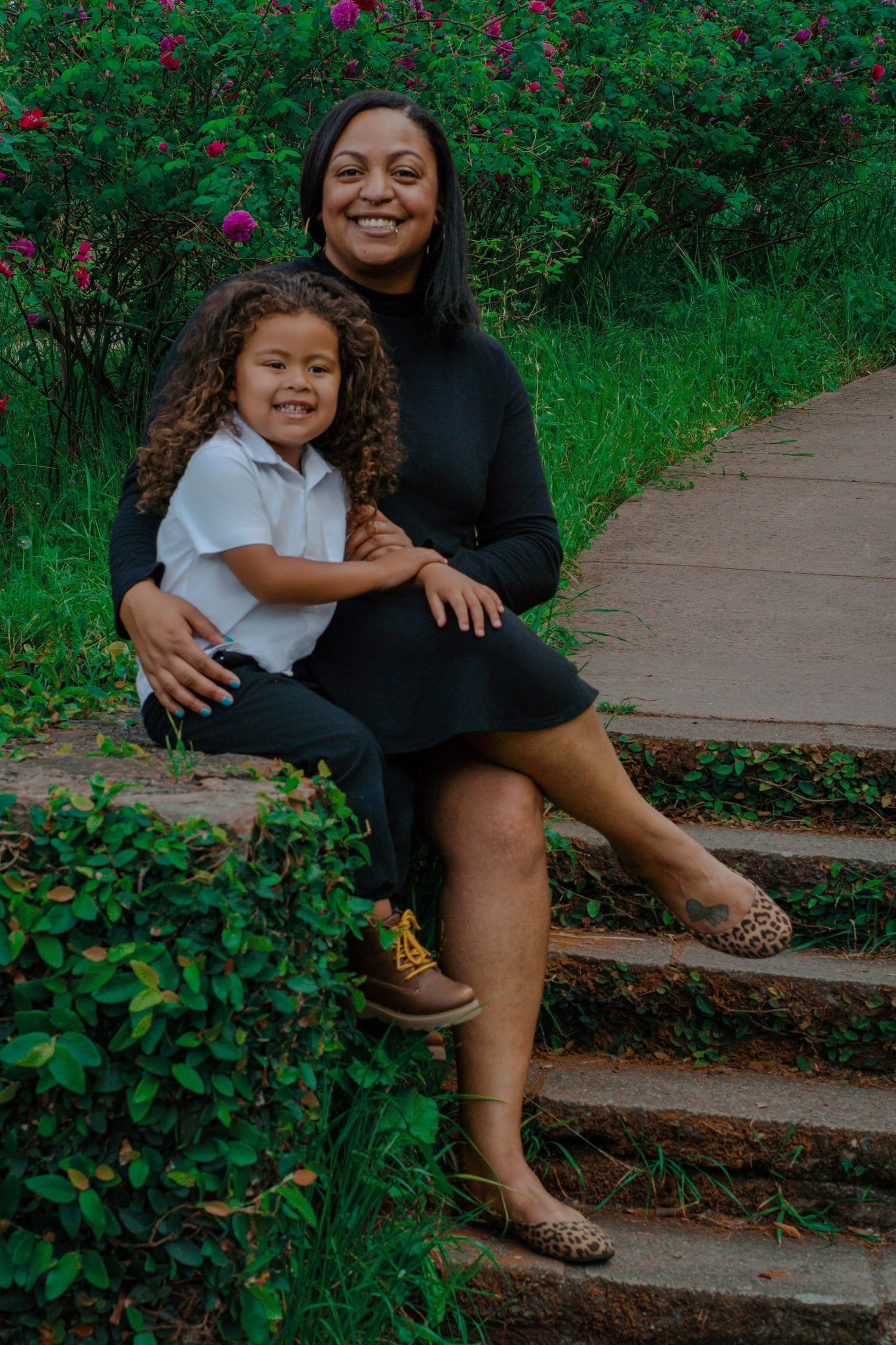 A smiling Black woman in a black dress sits on garden steps hugging a curly-haired child in a white polo.