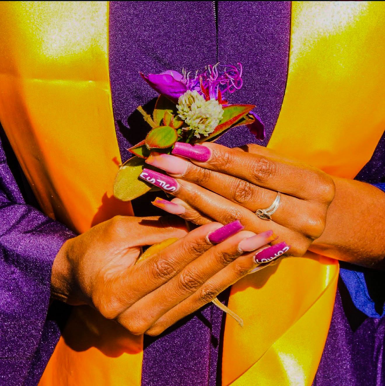 Close-up of hands with pink acrylic nails wearing a graduation gown in purple and gold with a floral boutonniere.