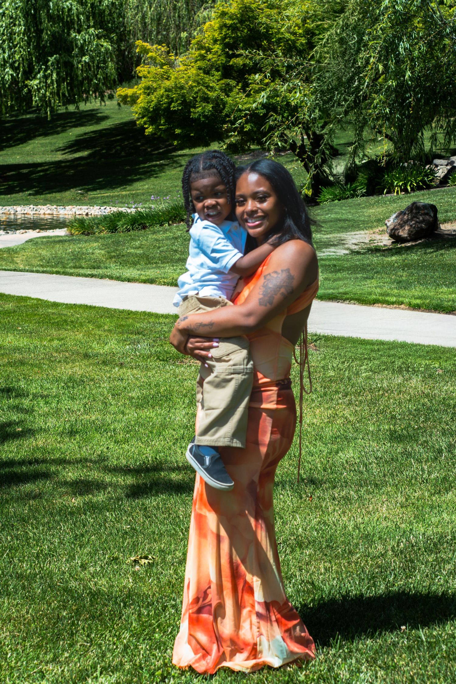 A smiling Black woman in an orange maxi dress holds a young child in a sunny green park.