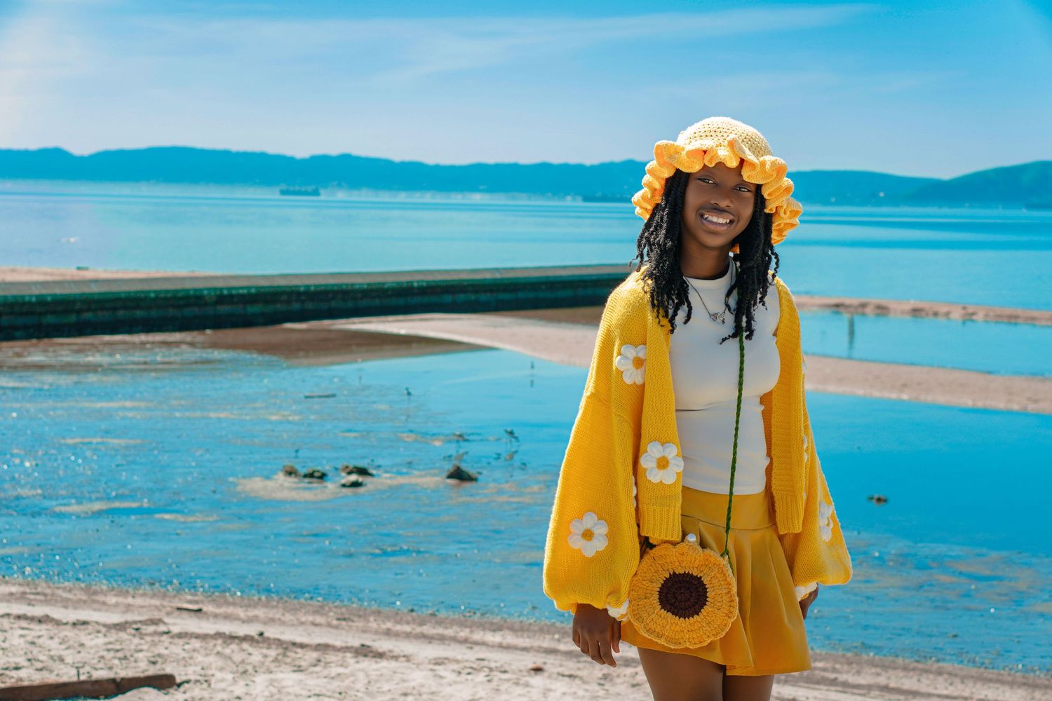 A smiling woman in a yellow floral outfit and hat poses on a sunny beach with blue water in the background.