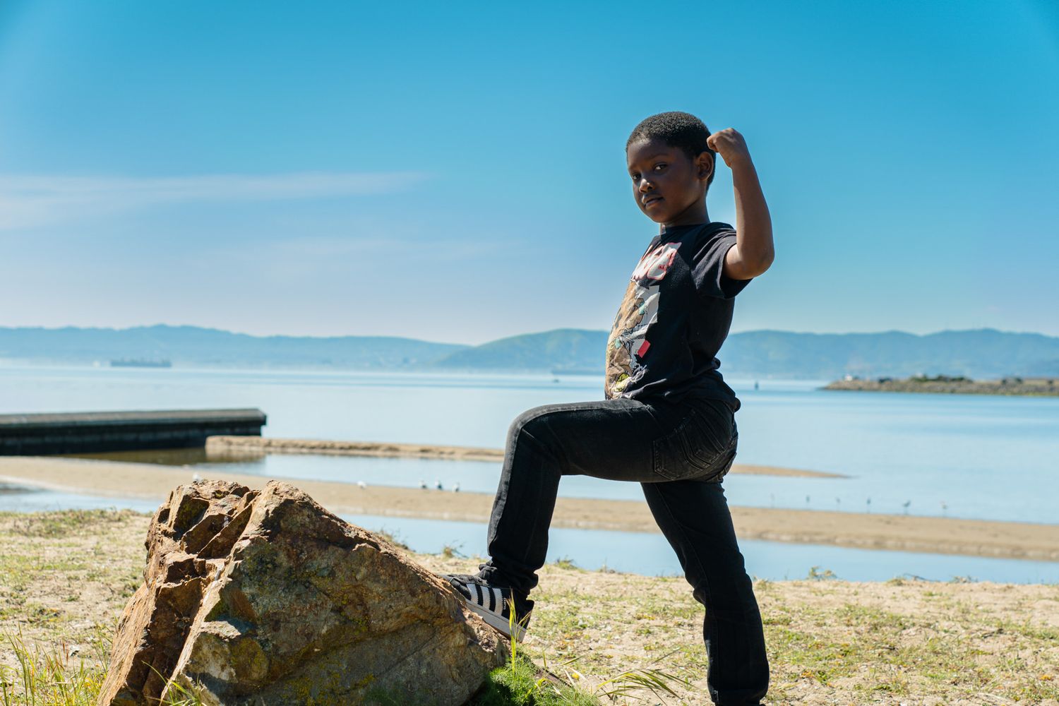 Young boy posing confidently on a rock with a scenic waterfront and blue sky behind him.