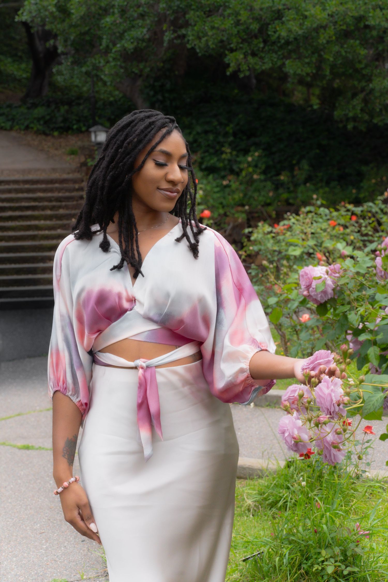 Woman in cropped tie-dye tie-front top and satin skirt touching pink roses in garden.