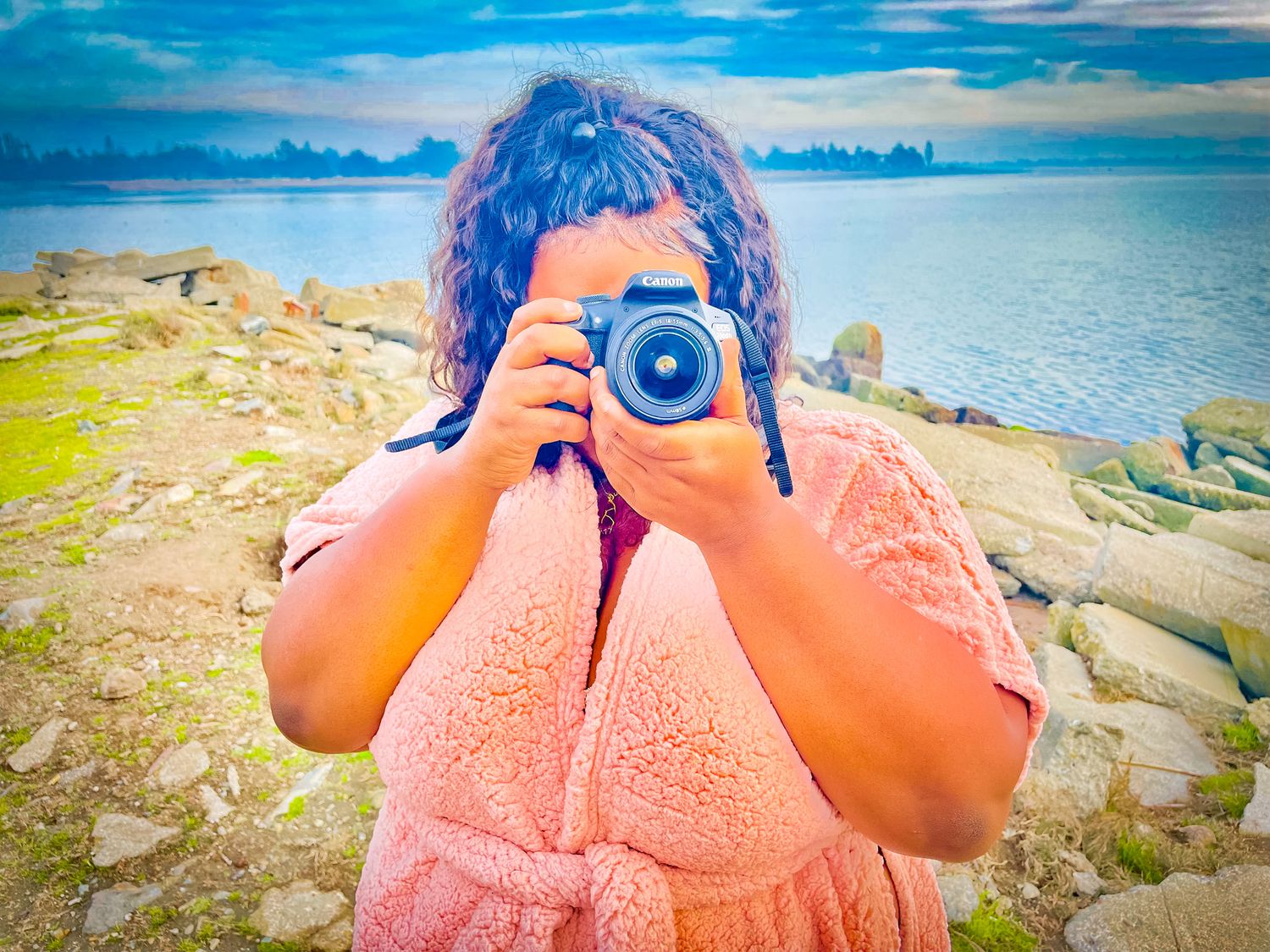 Woman photographing with DSLR camera outdoors on rocky coastal landscape with blue water and sky background.