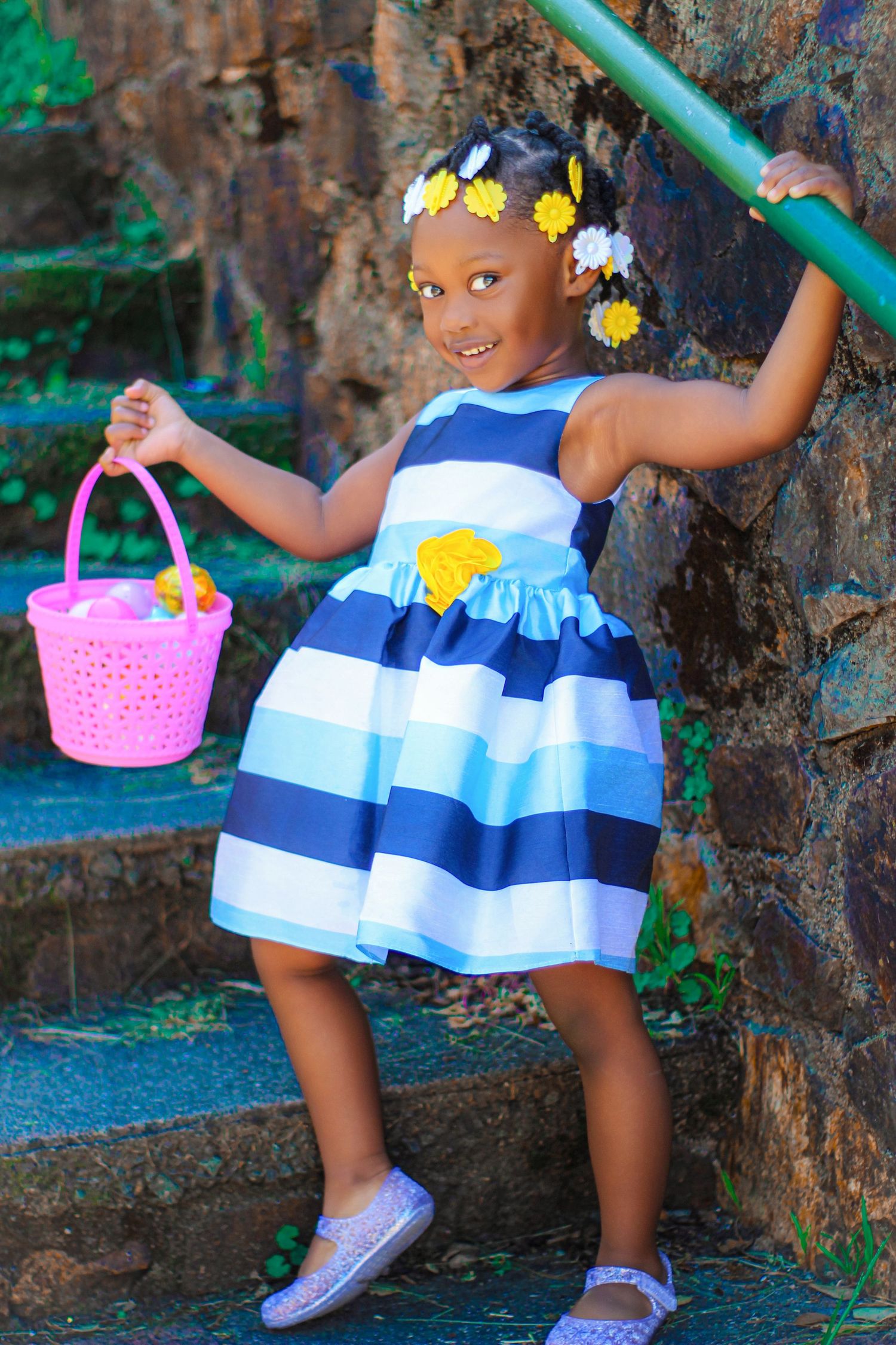 Young girl in striped blue dress holding pink Easter basket, wearing yellow flower hair accessories on stone steps.