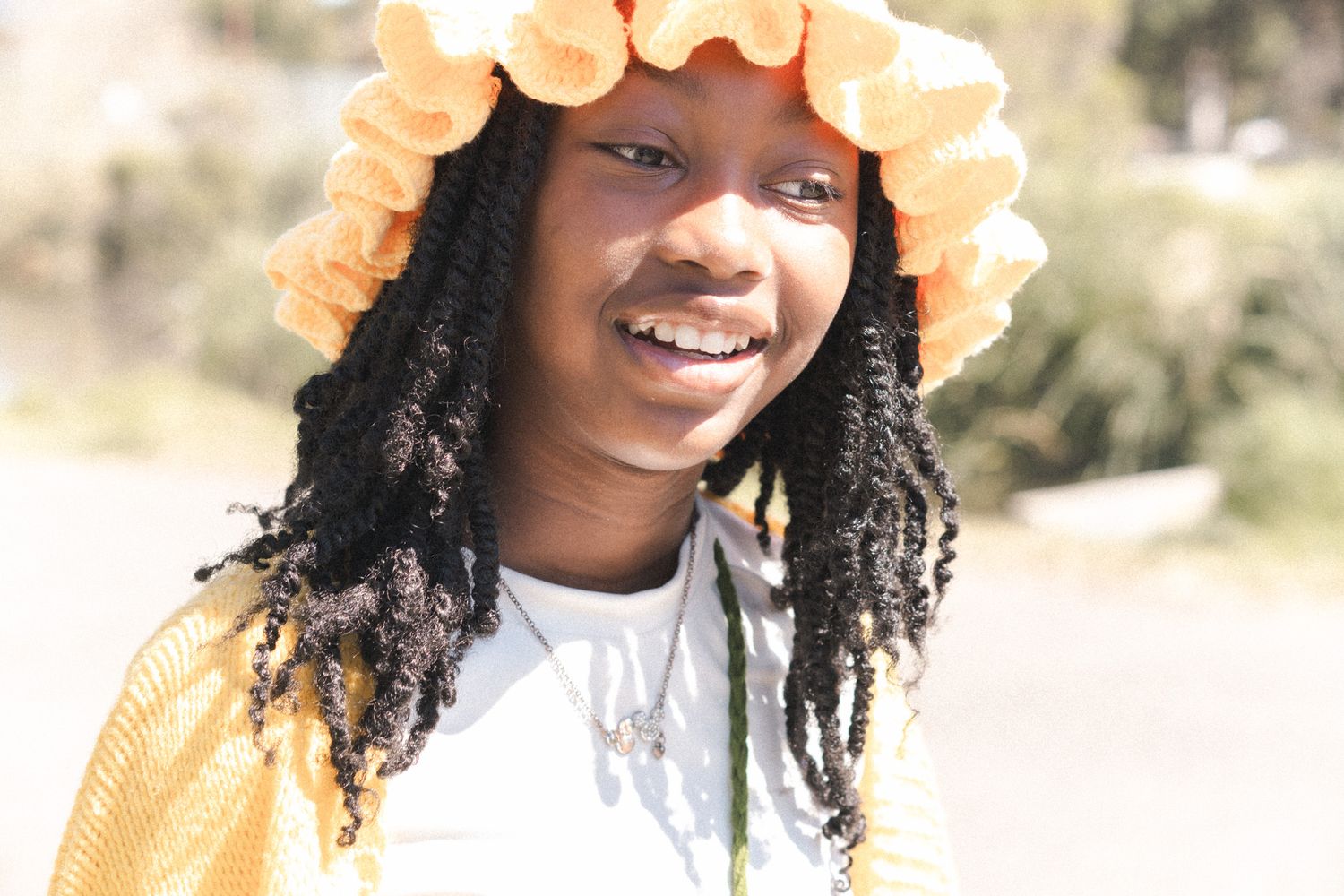 Smiling young girl wearing a yellow flower bucket hat and braids outdoors in sunlight.